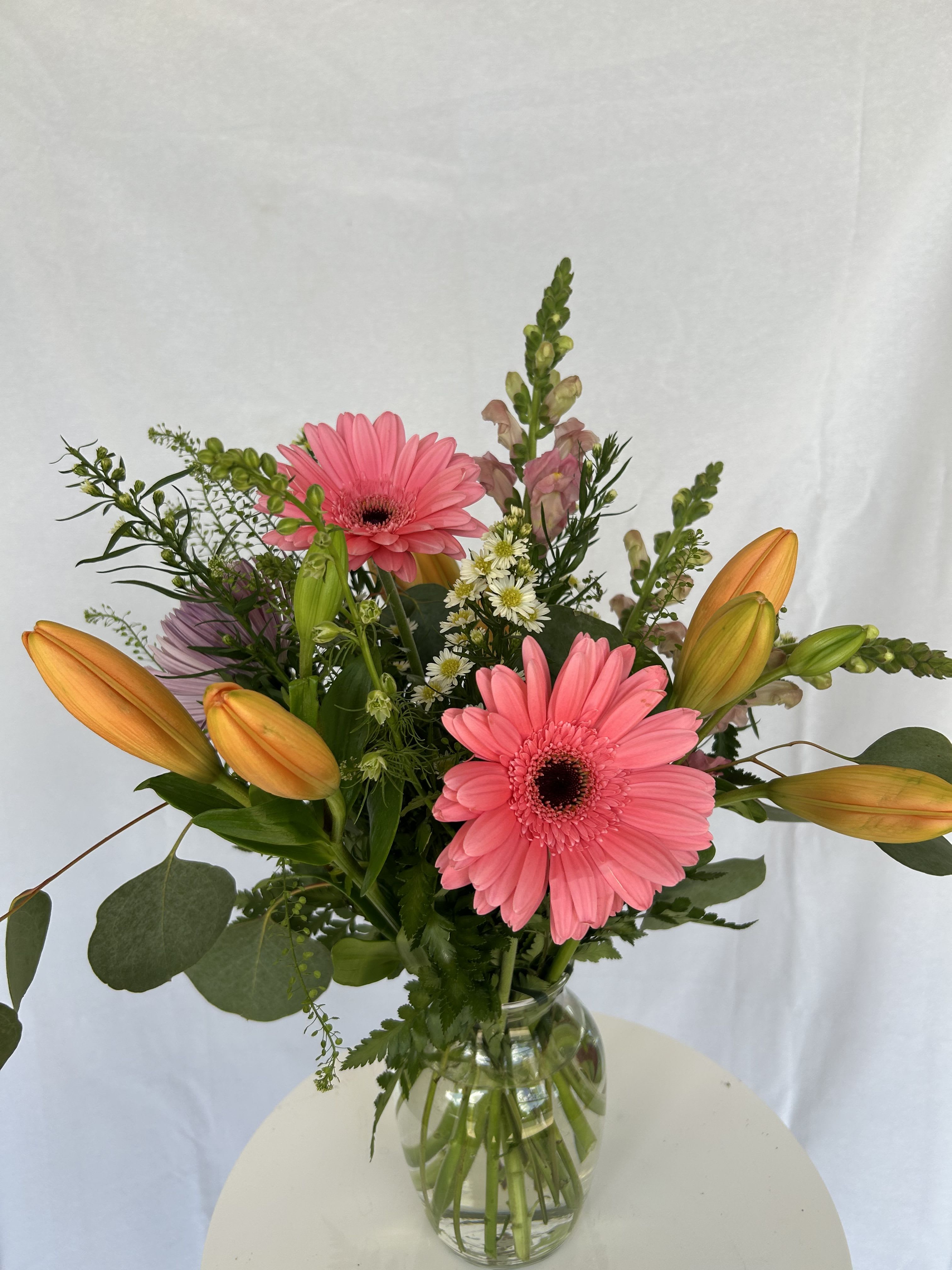 Sassy Stems  - A bright and cheerful arrangement that instantly lifts the mood. This design features bold hot pink gerbera daisies as the focal point, paired with vibrant orange lilies just beginning to bloom—promising even more beauty in the days ahead. Soft blush snapdragons and delicate white filler flowers add texture and movement, while fresh greenery brings everything together with a natural, garden-fresh feel. Designed in a clear glass vase, this arrangement is the perfect balance of playful and polished—ideal for birthdays, celebrations, or simply adding a burst of color to someone’s day.