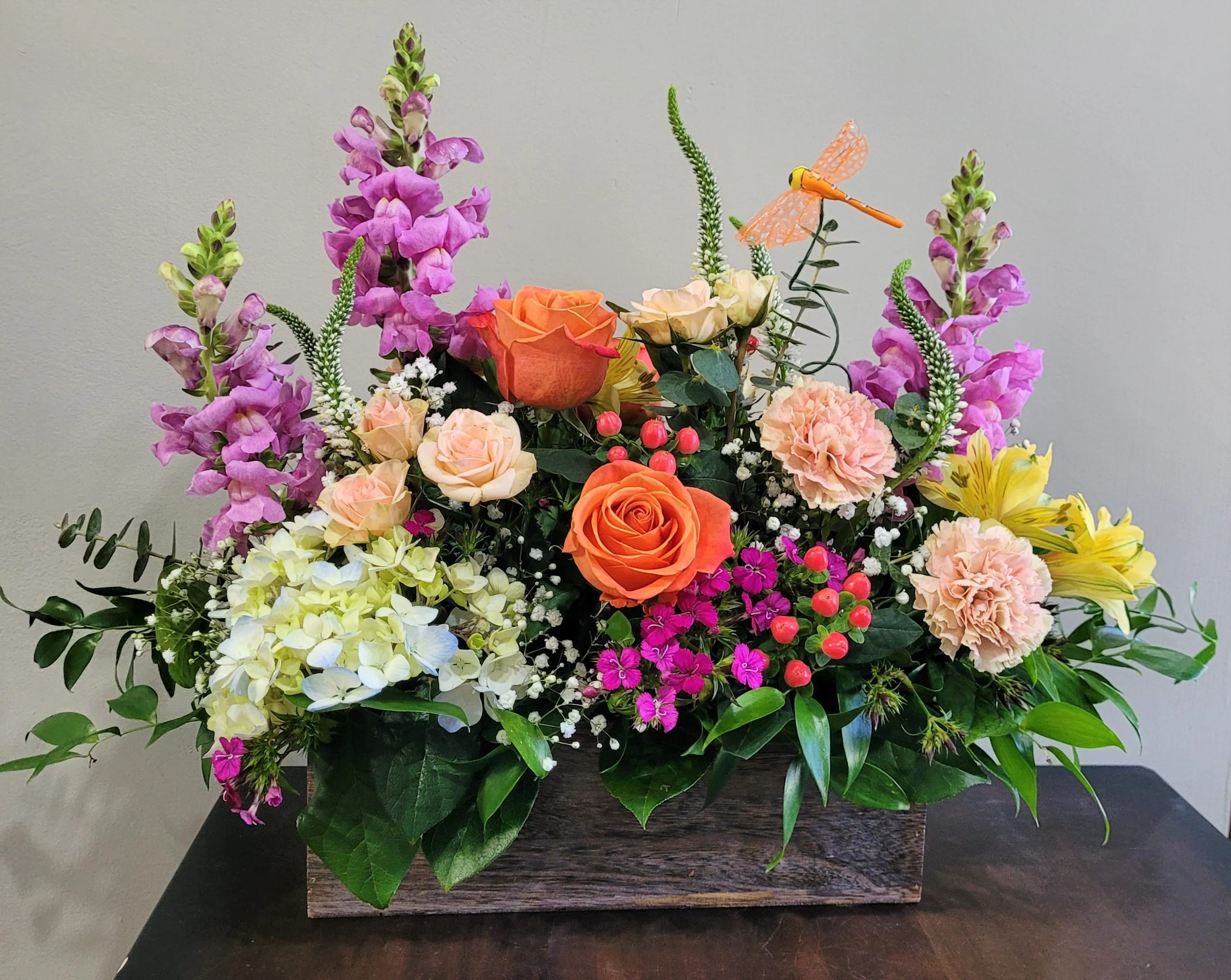 Rectangular wooden box filled with orange roses, pink carnations, purple snapdragons, and yellow blooms
