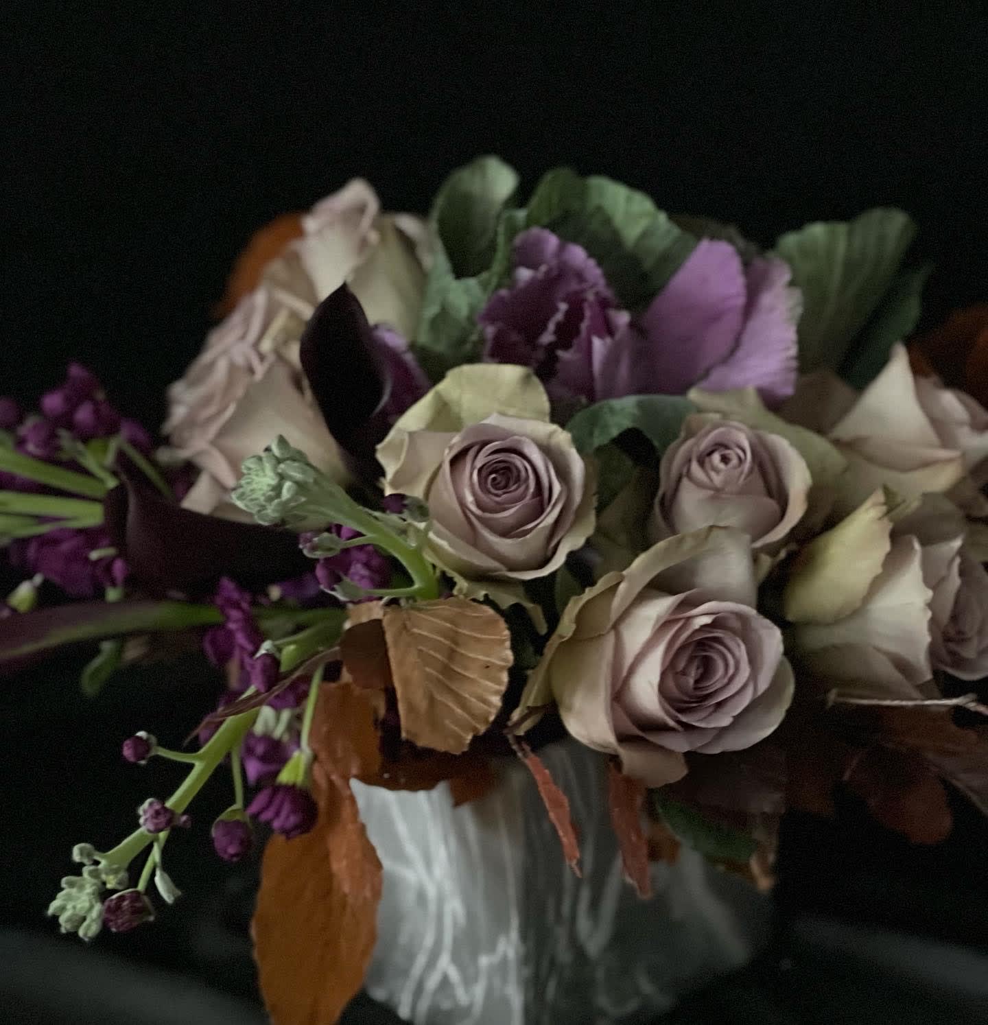 Moody arrangement of lavender roses, dark purple flowers, and kale in a white ceramic vase