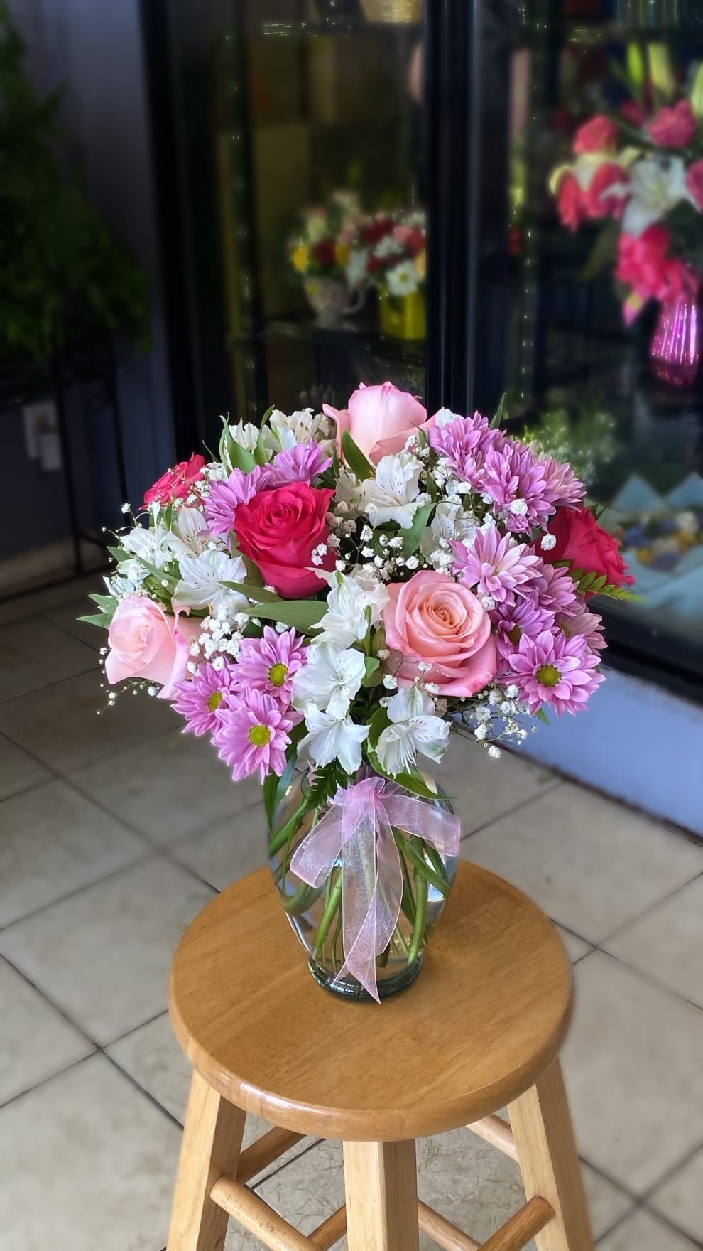 Pink roses, lavender daisies, and white blooms in a clear glass vase with a pink ribbon.