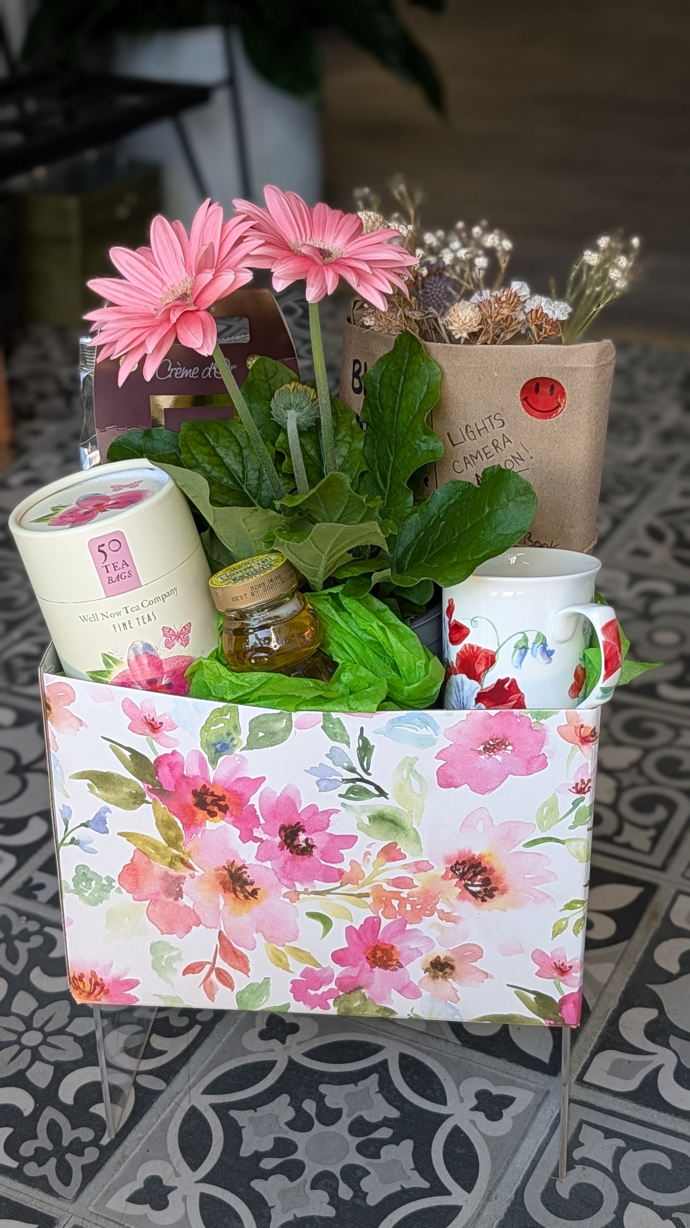 Gift box with a potted pink gerbera daisy plant, tea, honey, mug, and treats in a floral-patterned container