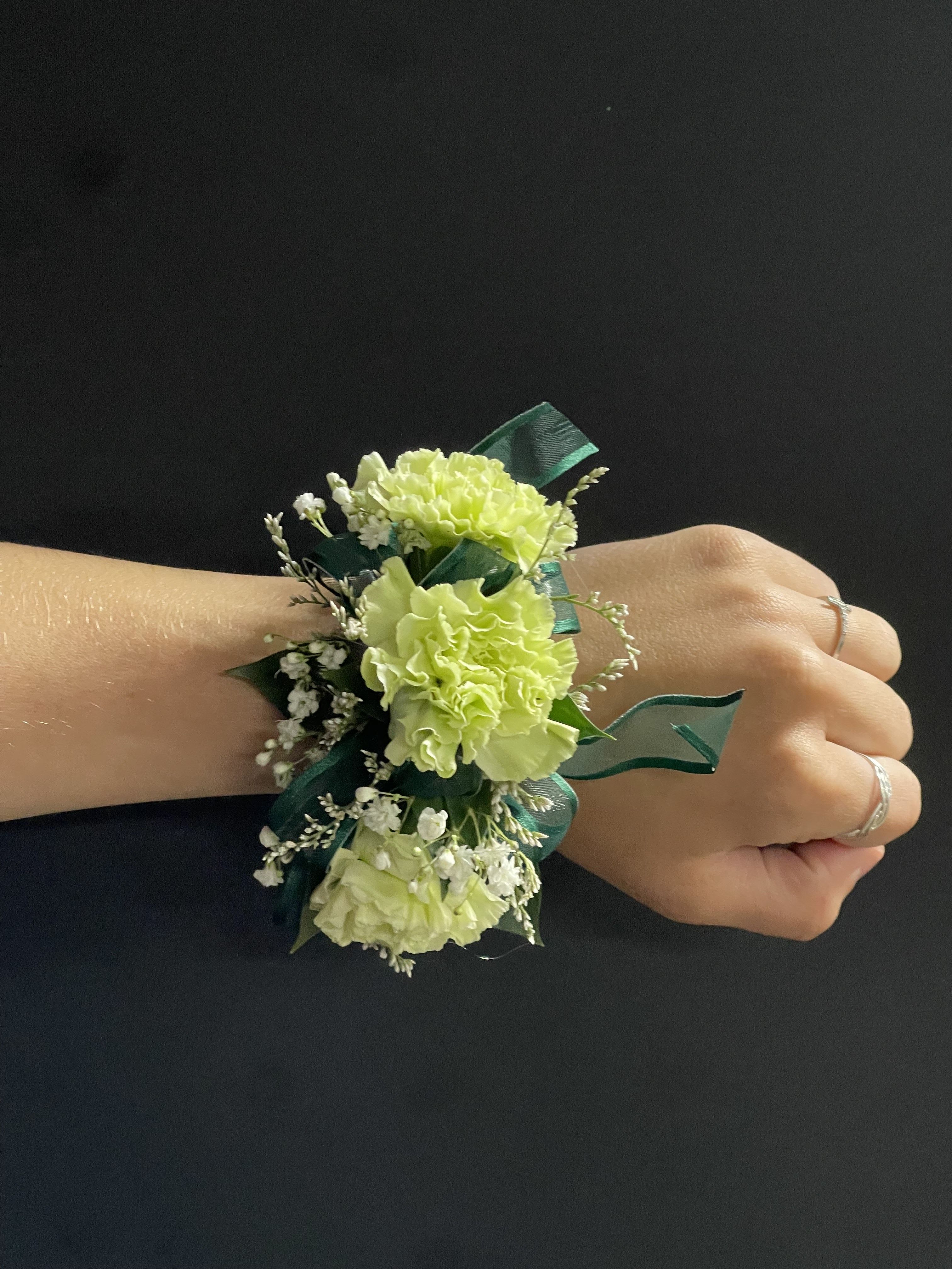 Light green carnation wrist corsage with white filler flowers and dark green ribbon on a hand.