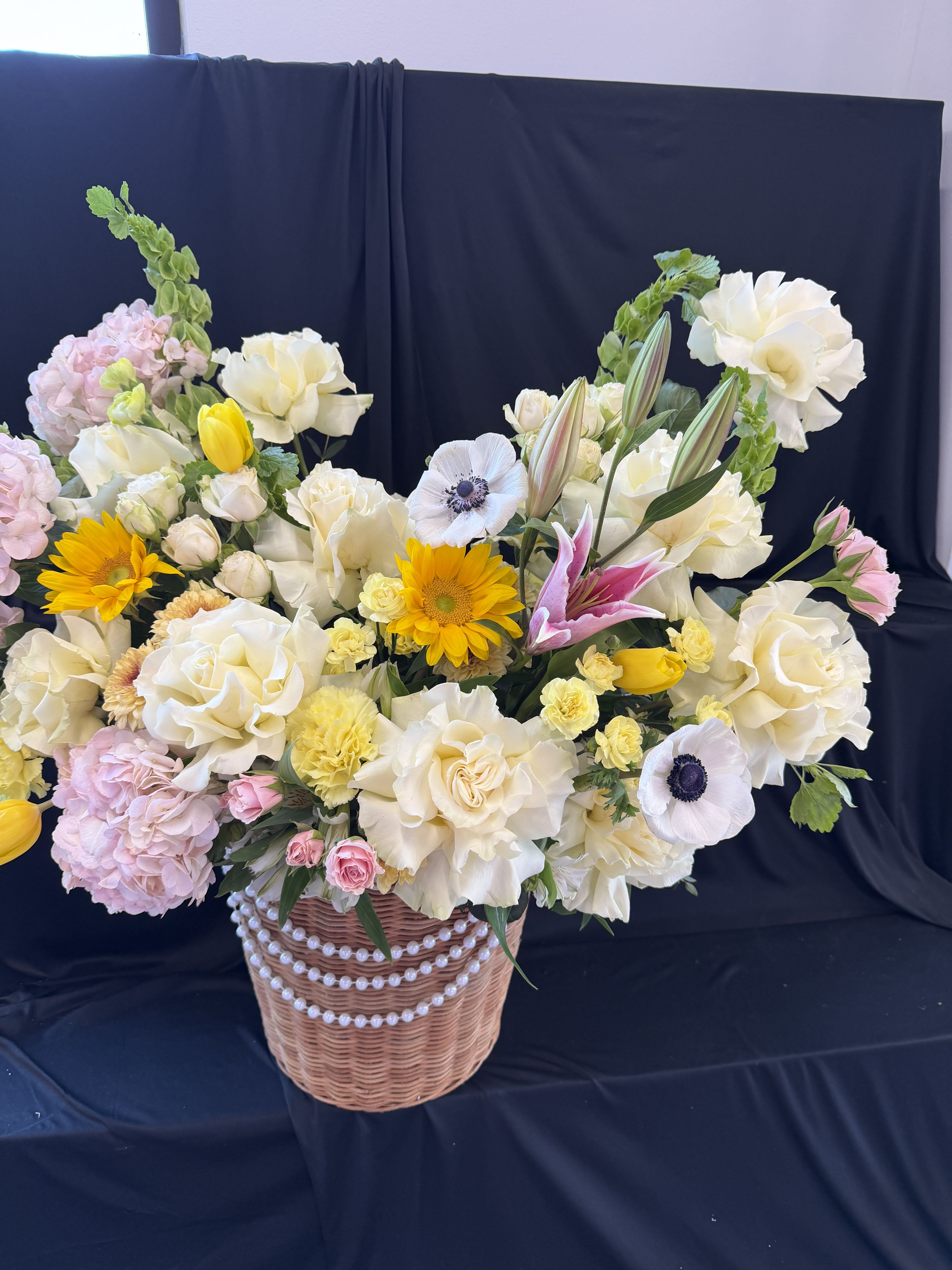 Golden Blush Garden - This charming basket arrangement features lush cream roses and ruffled ivory lisianthus, beautifully paired with soft blush hydrangeas for a romantic base. Bright pops of sunshine come from cheerful yellow sunflowers and delicate yellow tulips, adding warmth and energy. Elegant pink lilies and striking white anemones with dark centers bring a touch of sophistication, while accents of spray roses, mini carnations, and seasonal filler flowers create fullness and texture. The design is completed with fresh greenery and presented in a woven basket adorned with pearls for a graceful, timeless finish.