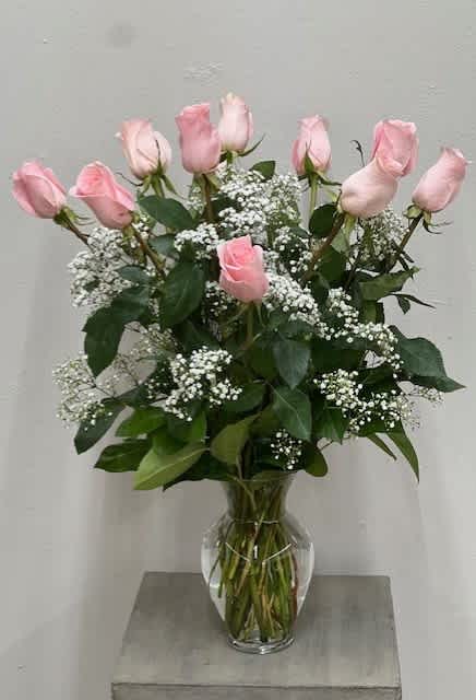 Tall clear vase of light pink roses with white filler flowers arranged in a rounded display