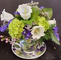 Compact arrangement of white and green flowers with purple accents in a patterned teacup and saucer