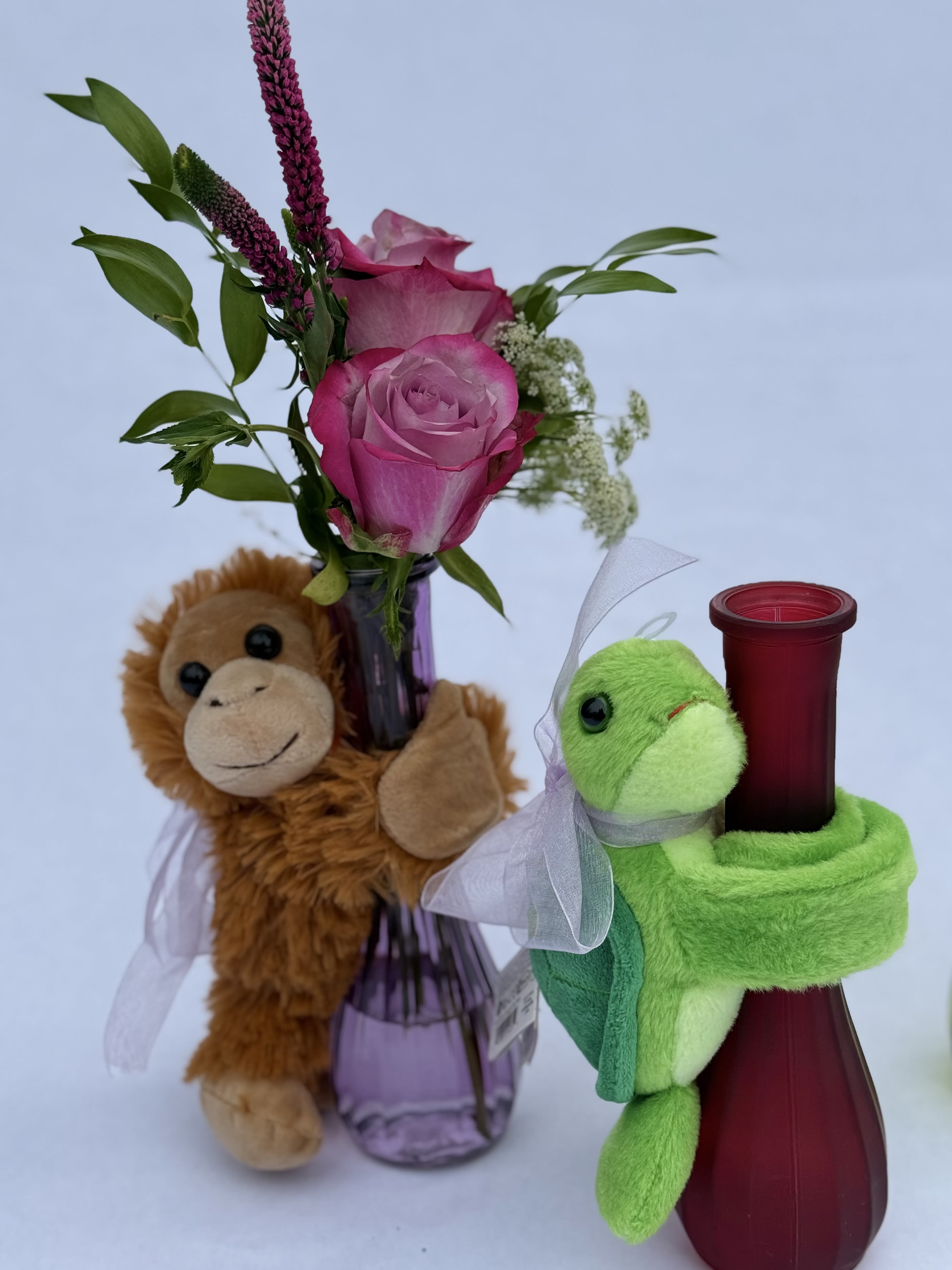 Pink roses in a purple bud vase with a monkey plush and a red vase hugged by a green turtle plush.