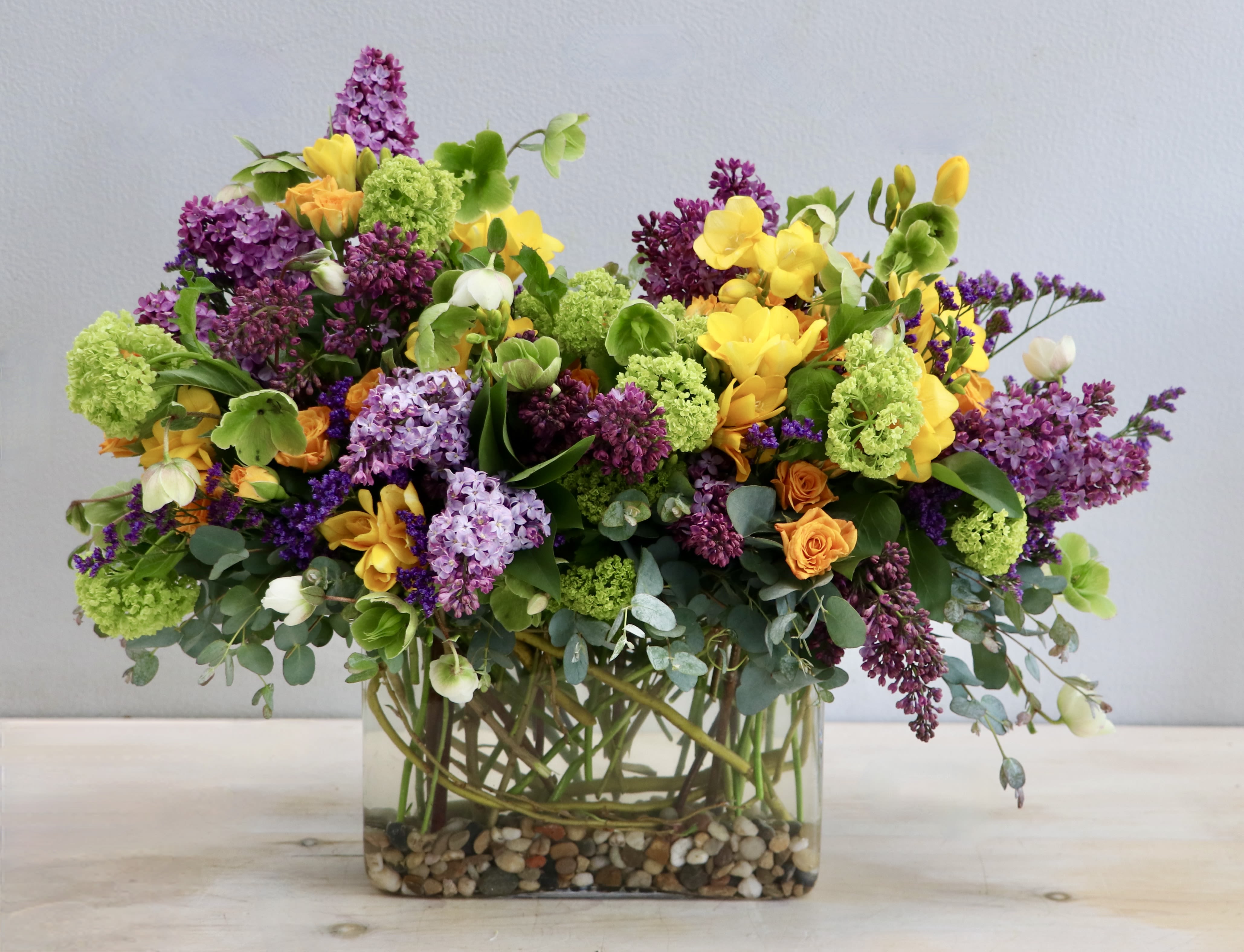 Lush mixed arrangement of purple, yellow, green, and orange flowers in a rectangular glass vase with pebbles.