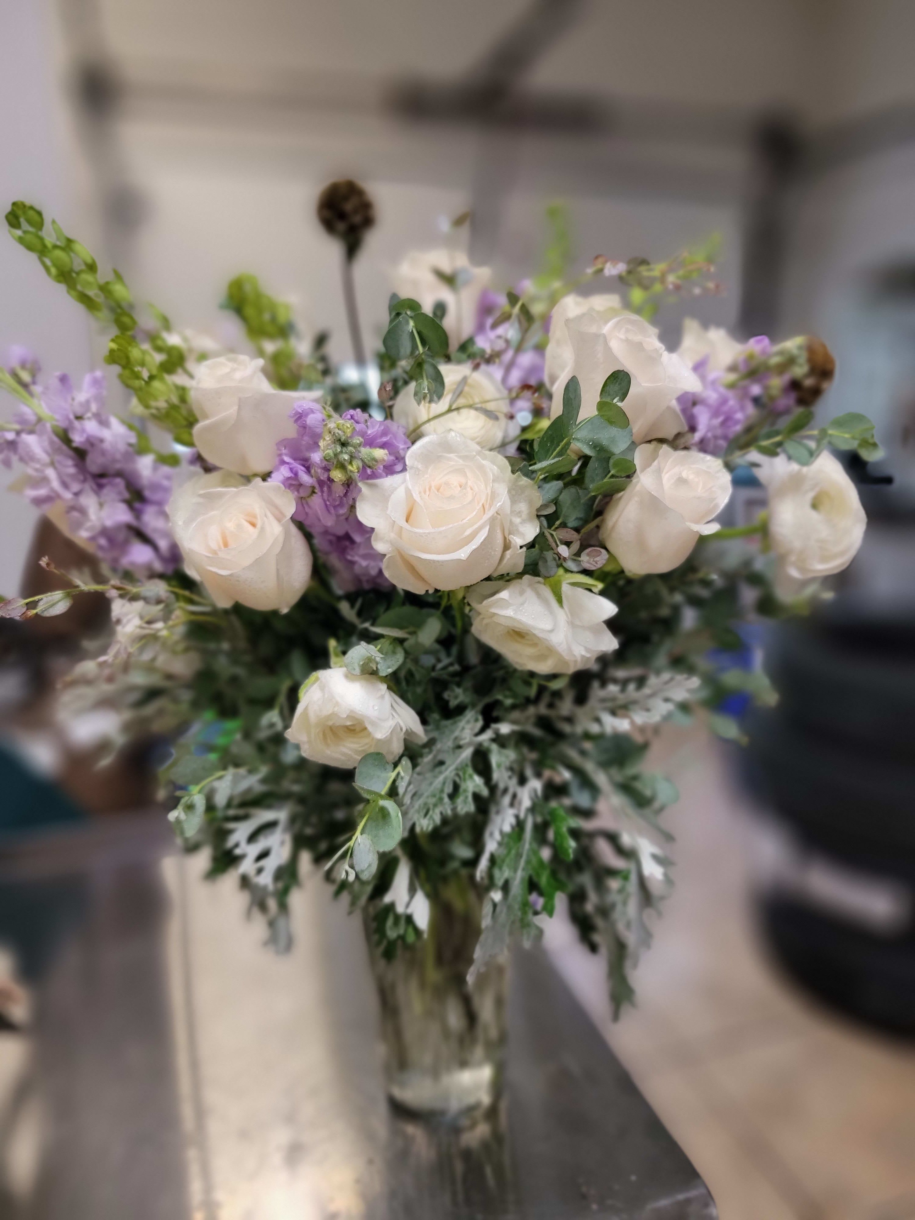 Elegant Rose Vase - Creamy white roses and soft lavender stock combine in a tranquil vase arrangement that reads as elegant and soothing-perfect as an anniversary bouquet, a get-well gift, or a sympathy arrangement. The tall clear glass vase showcases the stems for a refined, modern silhouette that lets the flowers take center stage and immediately conveys calm and care to the person you're honoring.  Clusters of white roses are woven with pale lavender stock for gentle contrast, while upright Bells of Ireland provide graceful height and a vertical rhythm above the main floral mass. Textural seed pods and silvery, lacy foliage at the base, plus scattered eucalyptus, add cool green accents and an airy finish that keeps the bouquet feeling light and composed. The clear glass vase emphasizes the arrangement's clean lines and makes an understated statement in any room.  Designed to comfort and celebrate, this arrangement is the kind of gift that makes someone feel truly seen-whether marking a milestone, wishing a speedy recovery, or offering sympathy. Vase-arranged for easy display, it arrives ready to enjoy; refresh the water every few days and trim stems as needed to keep blooms looking their best.