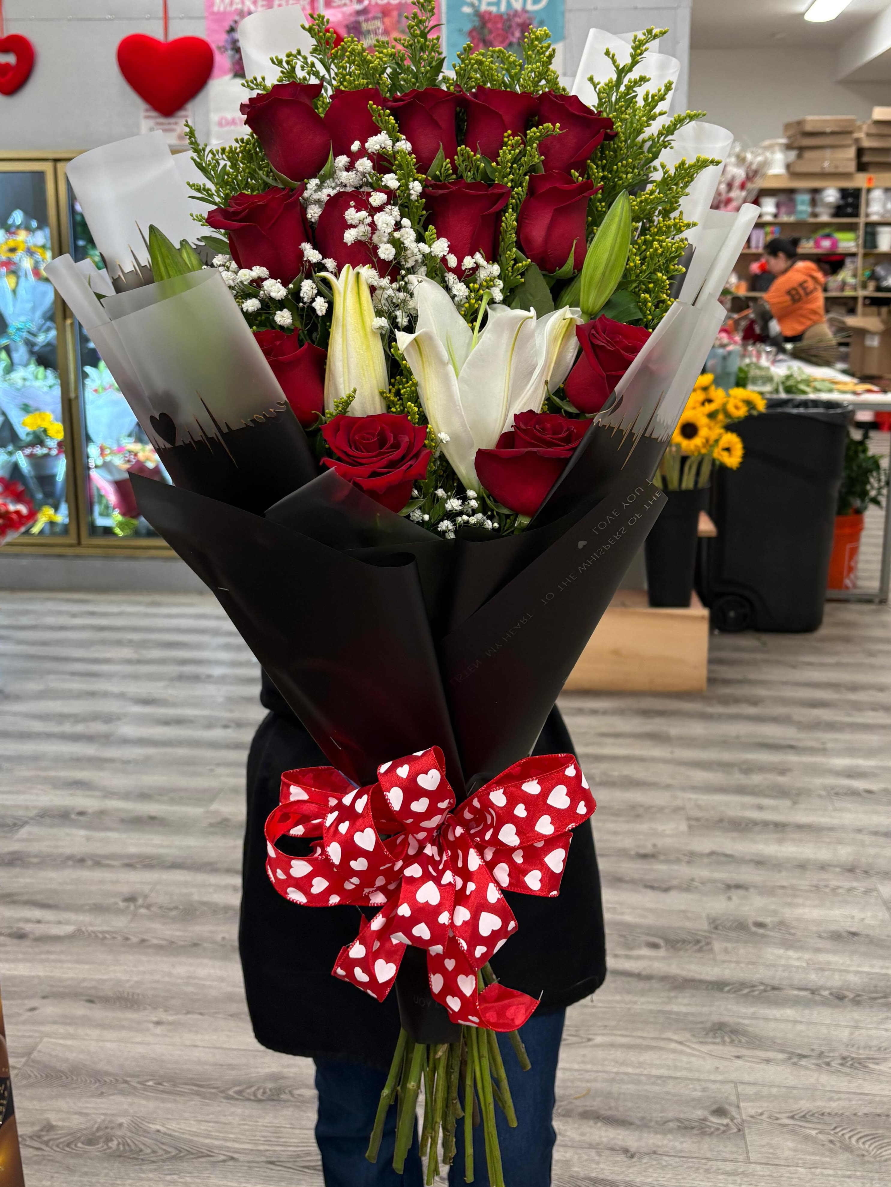 Tall wrapped bouquet of red roses and white lilies with a red heart-print bow