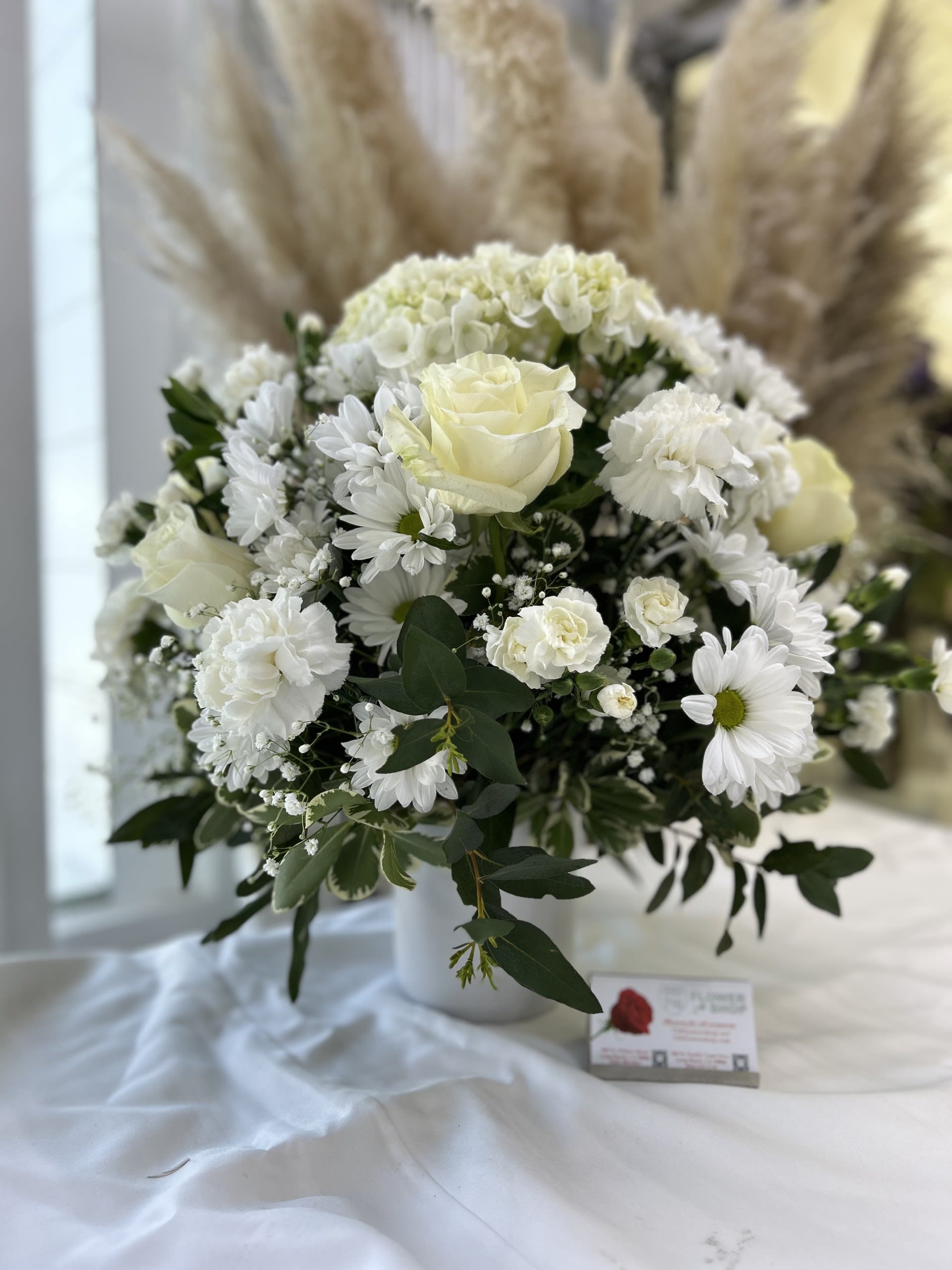 White roses, carnations, and daisies arranged in a white vase with feathery pampas stems in the background.