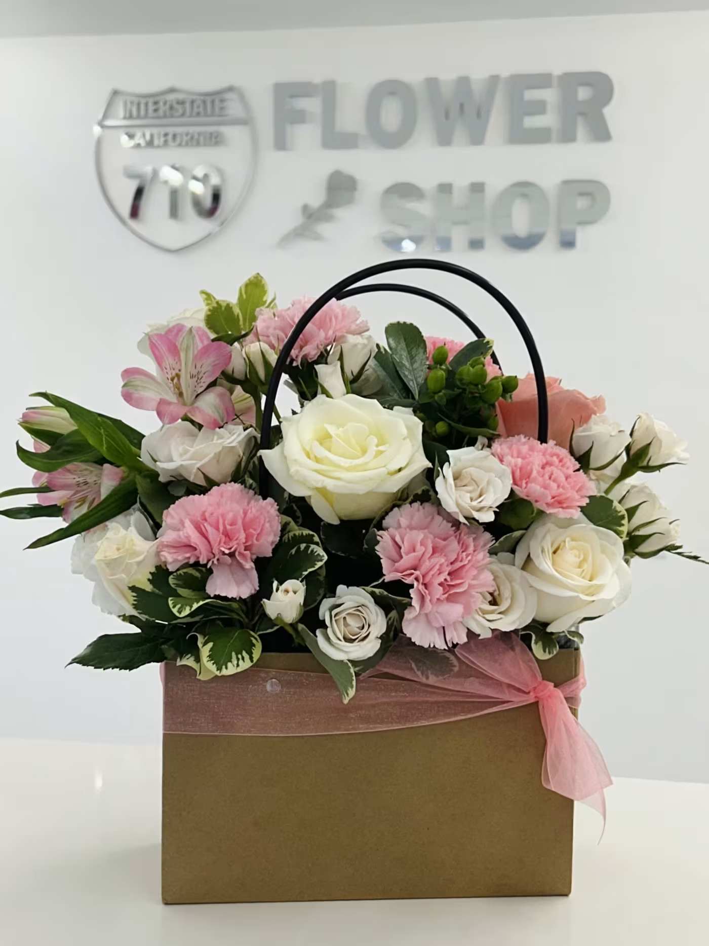 Garden Rose Delight Purse - A charming basket arrangement featuring a soft blend of blush pink and creamy ivory blooms, beautifully accented with fresh greenery and delicate textures. Designed in a handled basket and finished with a soft pink ribbon, this arrangement brings the gentle beauty of a blooming garden indoors. Perfect for birthdays, celebrations, thank-you gifts, or simply brightening someone’s day. (If some flowers and/or the vase are not available there will be minor substitutions.)