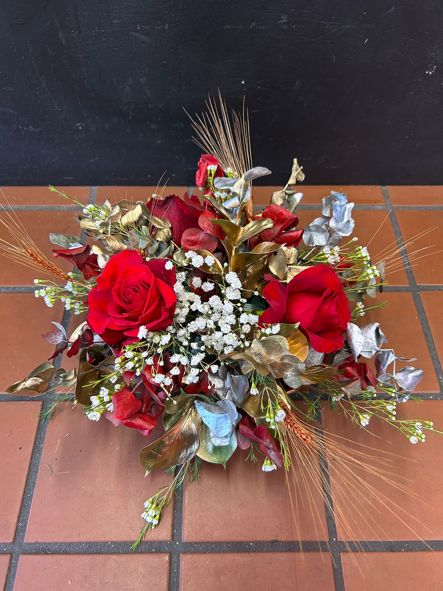 Low event centerpiece with red roses, white filler blooms, metallic foliage, and wheat accents.