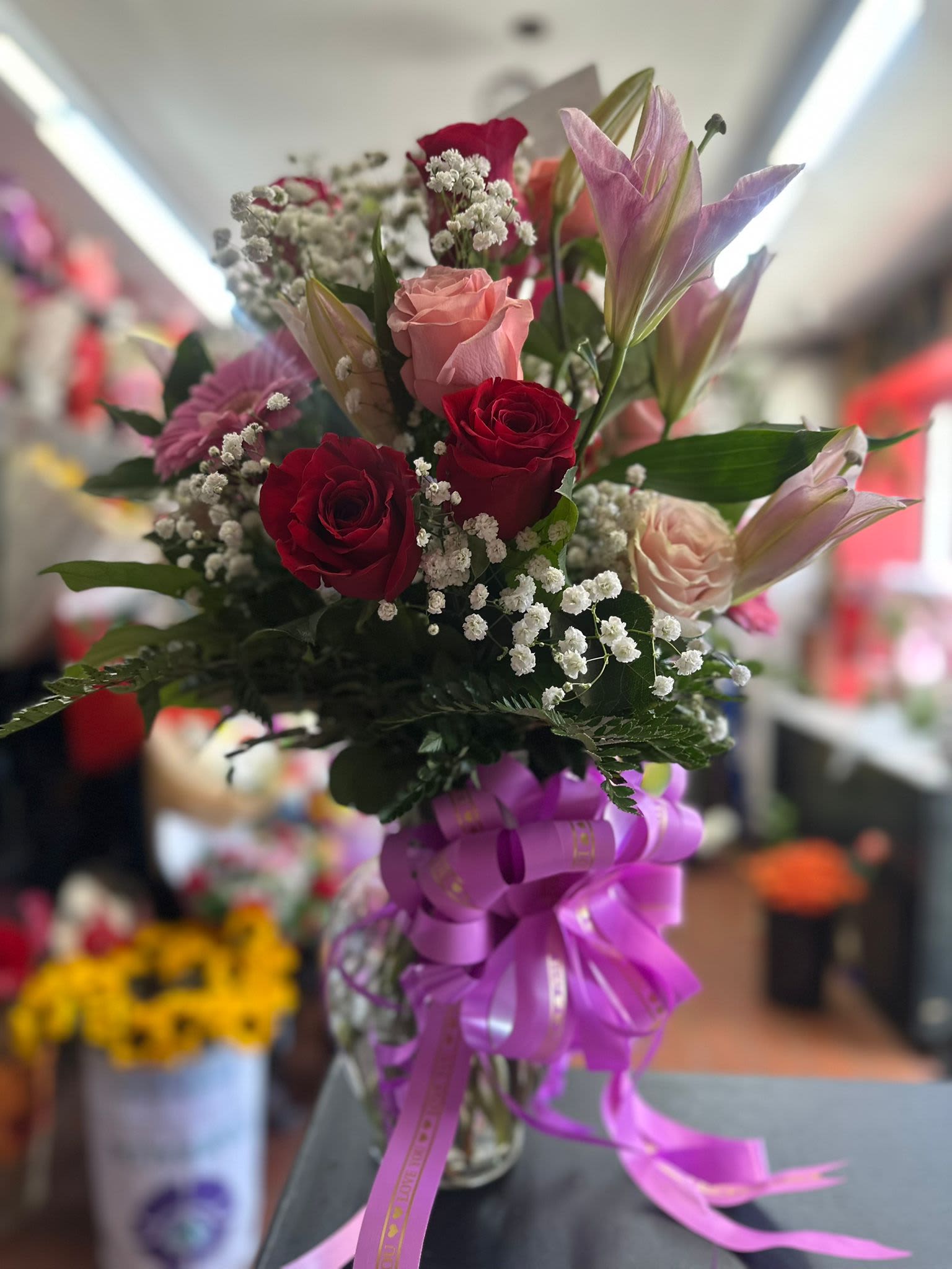 Tall vase arrangement of red and pink roses, pink lilies, and baby's breath with a large purple ribbon bow.