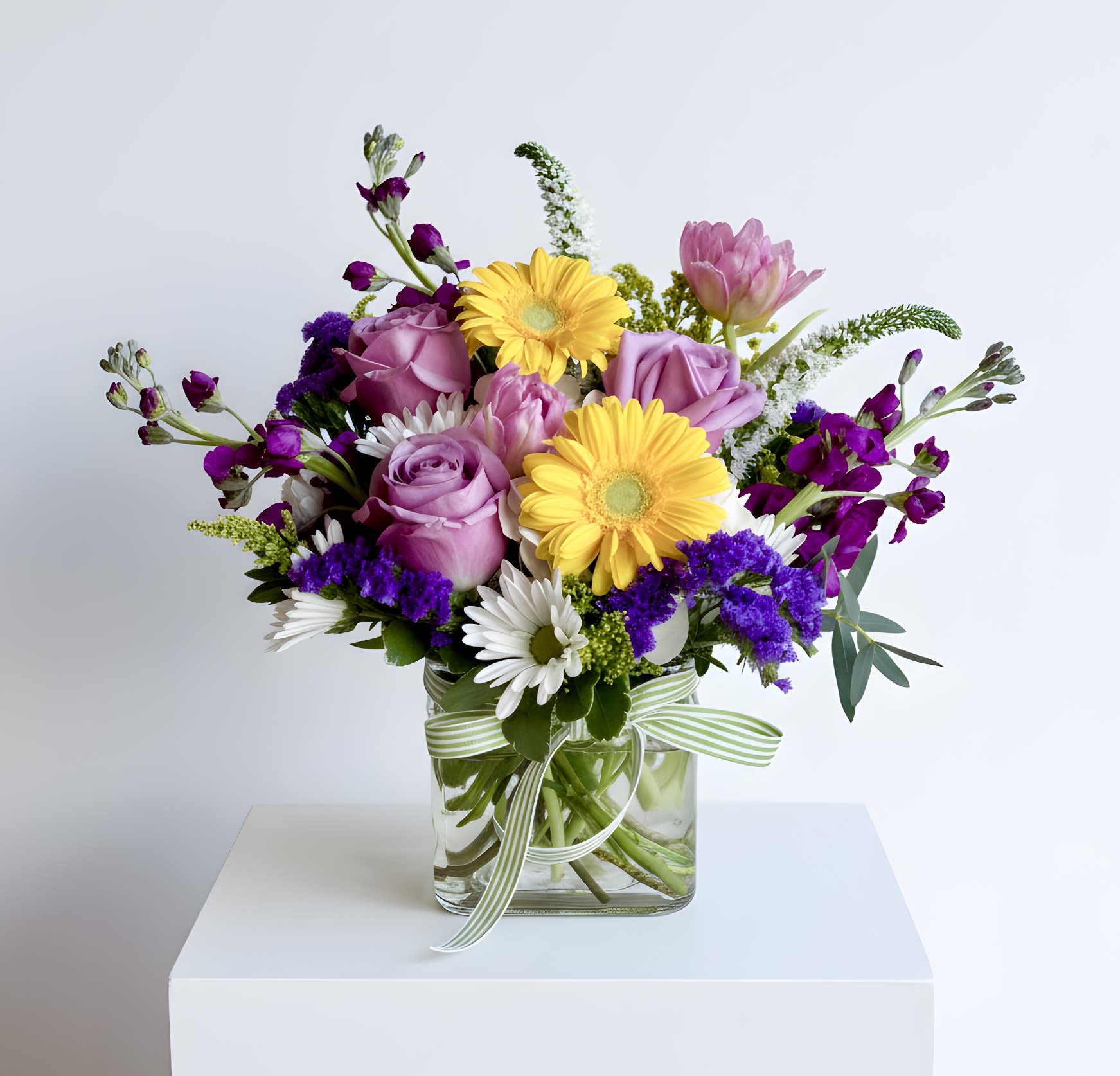 Mixed bouquet of purple roses, yellow gerberas and white daisies in a square glass vase with striped ribbon