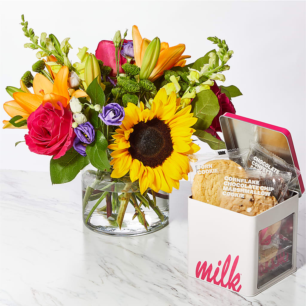 Bright mixed bouquet with sunflower, roses, and lilies beside a cookie tin on a marble surface
