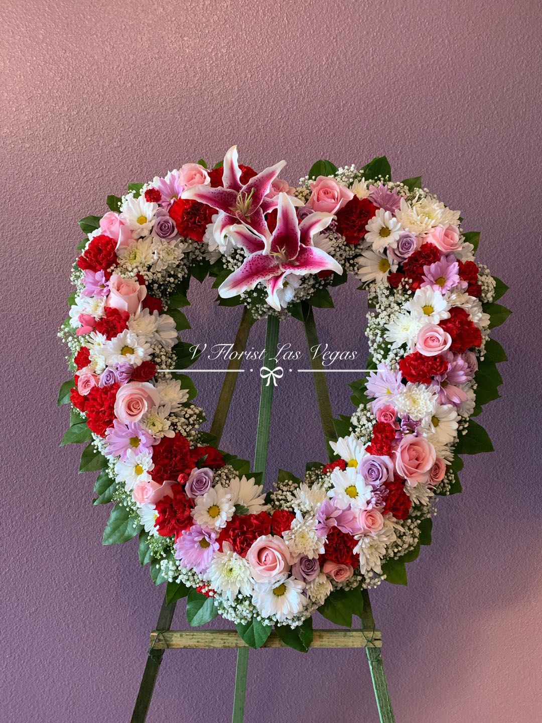 Heart-shaped standing wreath of pink lilies, roses, red carnations, and white daisies on an easel.