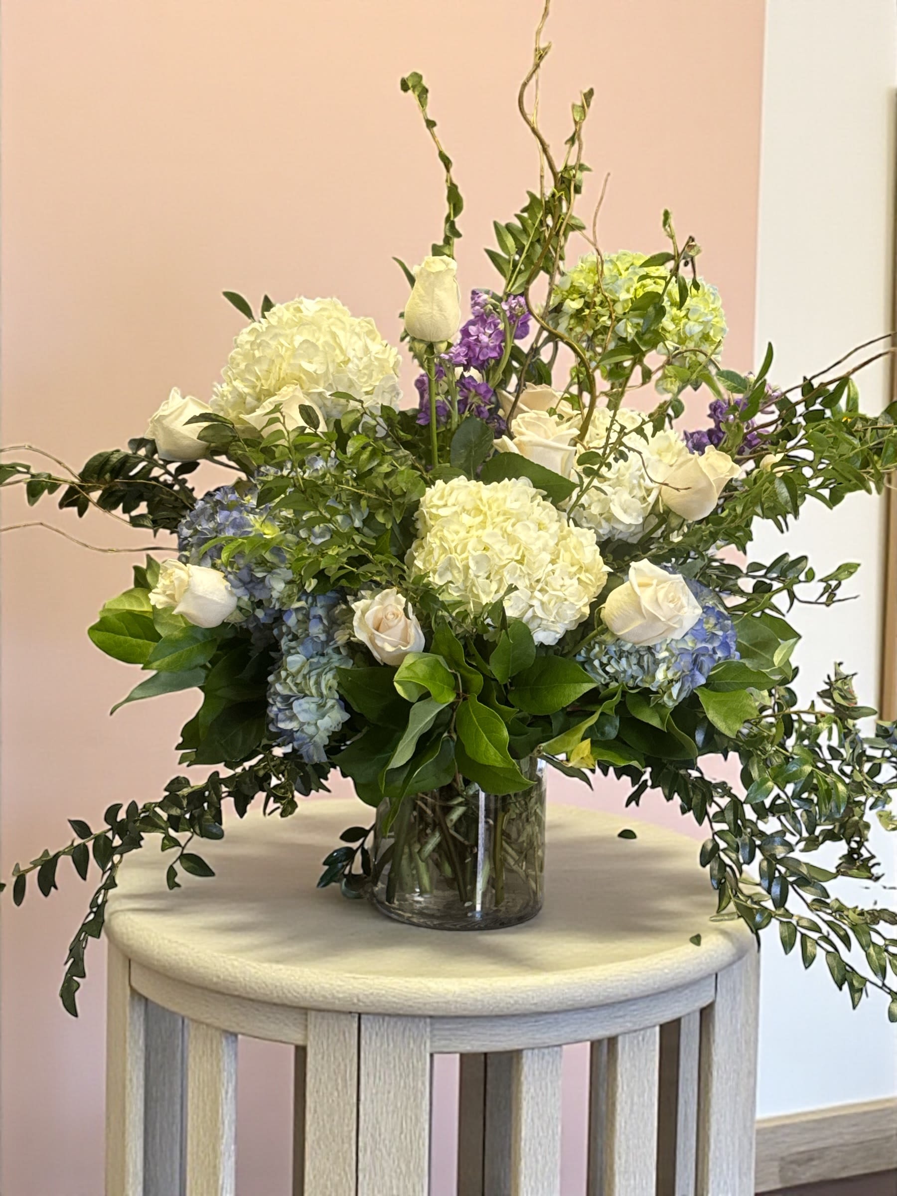 Garden Abundance - A lush, elegant floral arrangement displayed in a clear glass vase. The bouquet features large white hydrangeas as the focal blooms, accented by soft cream-colored roses and clusters of pale blue hydrangeas. Pops of light purple flowers add contrast and height, while abundant greenery surrounds and fills the arrangement, giving it a full, natural look.