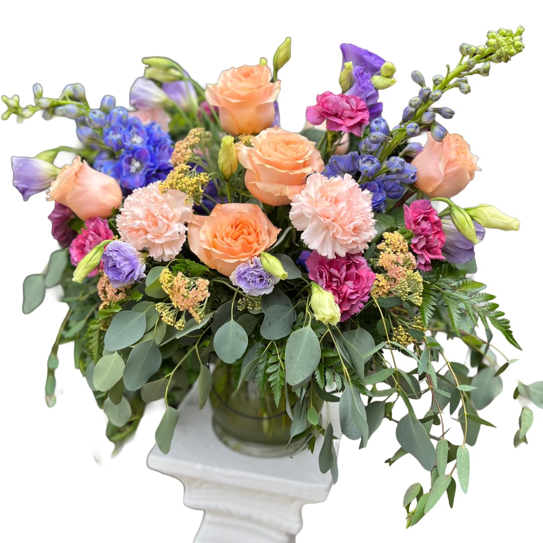 Pricilla - Peach roses and soft pink carnations take center stage in this airy arrangement, a joyful flower arrangement perfect for Mother's Day, anniversaries, birthdays, or simply to say "thinking of you." Purple lisianthus and deep blue delphinium add cool contrast while small yellow-toned filler blooms give texture and a sunlit lift-together they create a warm, uplifting bouquet that reads as both romantic and comfortingly cheerful.  Arranged in a clear glass vase and set on a white pedestal stand, the blooms are gathered with abundant trailing foliage that spills outward, giving the arrangement a generous, garden-fresh silhouette. The mix of carnations, roses, lisianthus, delphinium, and delicate fillers offers varied shapes and soft, touchable textures in peach, pink, purple, and blue-ideal for spring and summer celebrations or to brighten a bedside or entry table.  This is the kind of gift that makes the recipient feel truly seen-perfect for housewarming, get well, thank you, "I'm sorry," or just because moments when you want to lift someone's spirits. Available in a single vase size as shown; fresh-cut stems arrive ready to display-trim and refresh the water every few days to prolong bloom life.