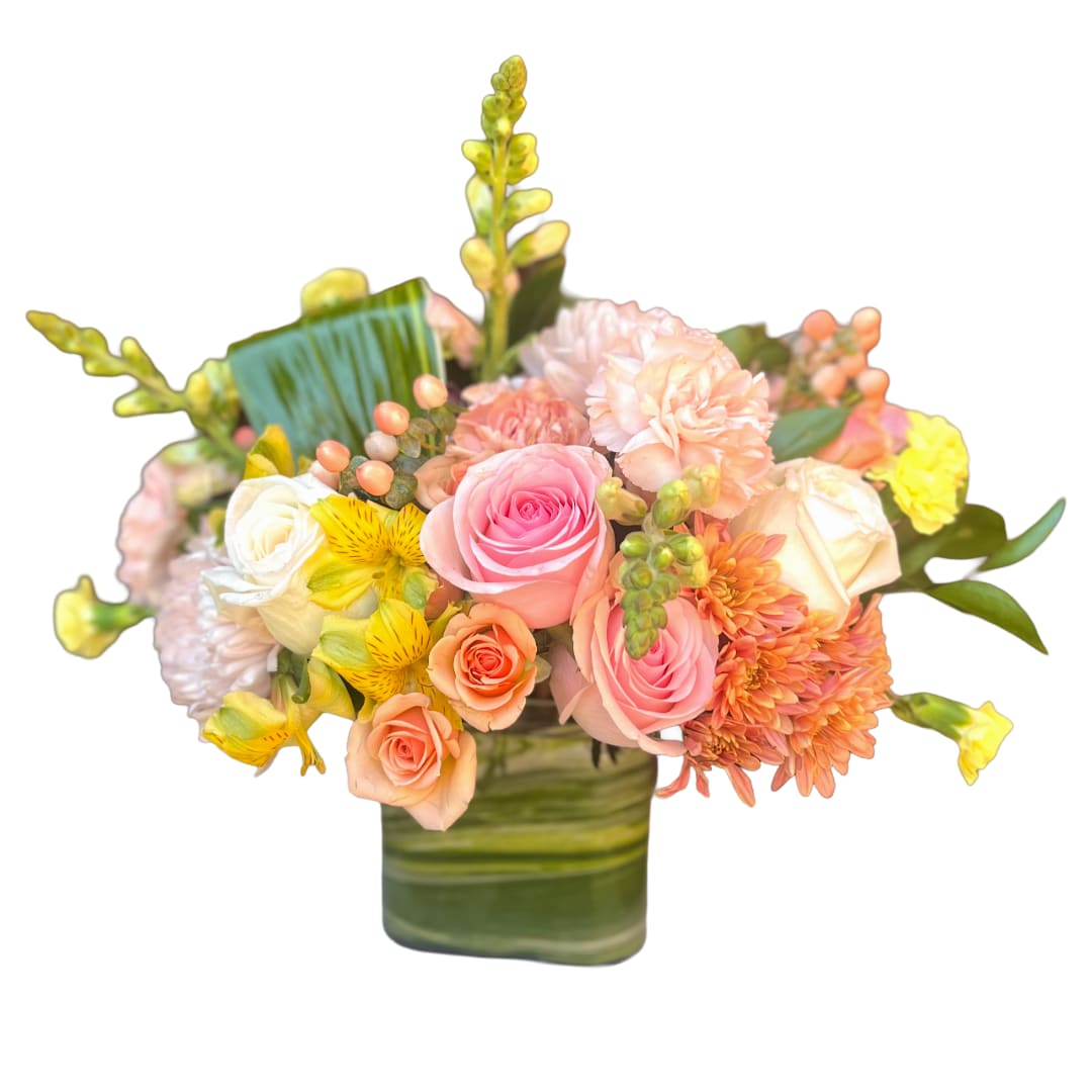 Marmalade - Soft peach and pink roses take center stage in this cheerful celebration bouquet-perfect as a birthday bouquet, get-well gift, or a heartfelt "thank you" arrangement. The mix of roses, peach carnations, and fluffy chrysanthemums creates a warm, pastel palette that feels both gentle and uplifting, while bright yellow alstroemeria and small yellow blooms add sunny contrast to brighten someone's day.  Upright snapdragons add height and sculptural texture, giving the design a lively silhouette, and a square glass vase keeps the composition modern and neat. Broad green leaves line the cube vase for a polished presentation; colors include pink, peach, yellow, and green. This compact arrangement sends a clear message of care and cheer-ideal for the person you want to make feel genuinely seen and uplifted.