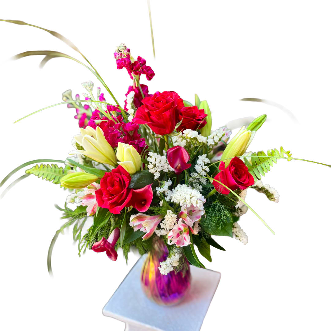 Lily Rose - Bold red roses meet pale yellow lilies in a romantic, celebratory arrangement made for anniversaries, birthdays, or simply to say "I'm thinking of you." The Lily Rose bouquet pairs the classic drama of roses and lilies with a lively mix of deep magenta stock and delicate pink and white alstroemeria, creating a layered, textured silhouette that feels both elegant and slightly wild.  Clusters of mini calla lilies and white filler blooms weave through the design, while tall blades of ornamental grass and arching fern fronds lift the composition for an airy, sculptural profile. The flowers are gathered in an iridescent glass vase that catches pink and gold light, giving the gift a luminous presence that will be noticed as soon as it's placed in a room.  This is the kind of arrangement that makes the recipient feel truly celebrated-romantic enough for an anniversary, joyful for a birthday, and thoughtful for a thank-you. Arrange on a tabletop or mantel; replace vase water every two days and trim stems slightly when refreshing to extend vase life.