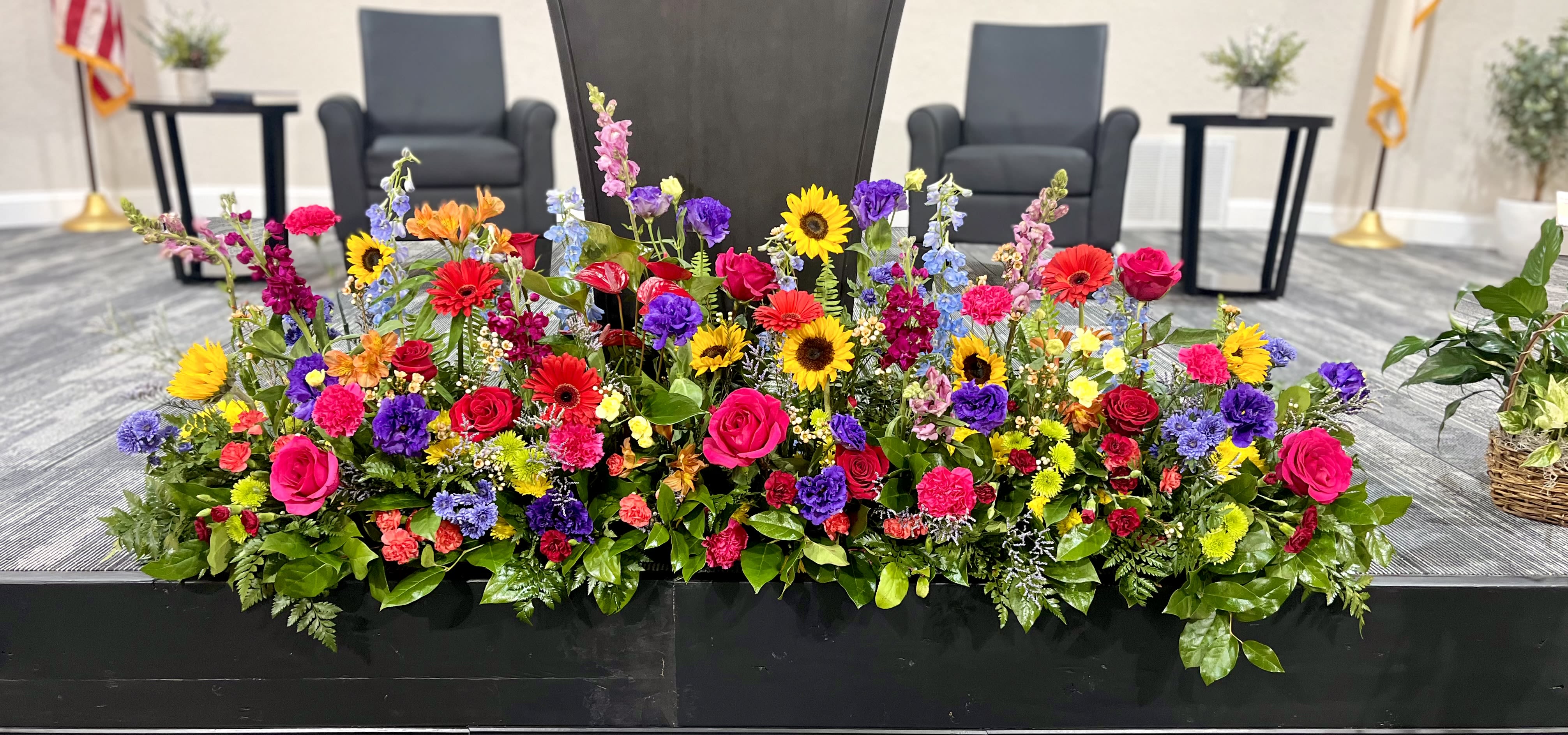 Isabelle’s Garden - Bright sunflowers and hot pink and red roses create an immediate burst of warmth and joy in this wide, garden-style flower arrangement-perfect as a graduation centerpiece, a vibrant wedding accent, or a colorful sympathy display for a celebration of life. Isabelle's Garden pairs bold yellow, pink, and red focal blooms with cooler purple and blue touches to give the person you're honoring a radiant, uplifting presence at any ceremony or gathering.  Designed low and wide in a sleek black rectangular container, the arrangement mixes sunflowers, gerbera daisies, roses, carnations, chrysanthemums, snapdragons, and delphinium for texture and movement. Varied heights and loose, natural placement mimic a freshly gathered garden; clusters of smaller yellow and green blooms add depth and continuity across the composition.  For seasonal spring events or year-round celebrations, this piece reads both joyful and respectful-she'll feel truly seen whether you're congratulating a grad, marking a wedding milestone, or offering comfort at a memorial. The low profile makes it ideal for long tables, stages, or service fronts; refresh water daily and keep stems trimmed to prolong bloom life.