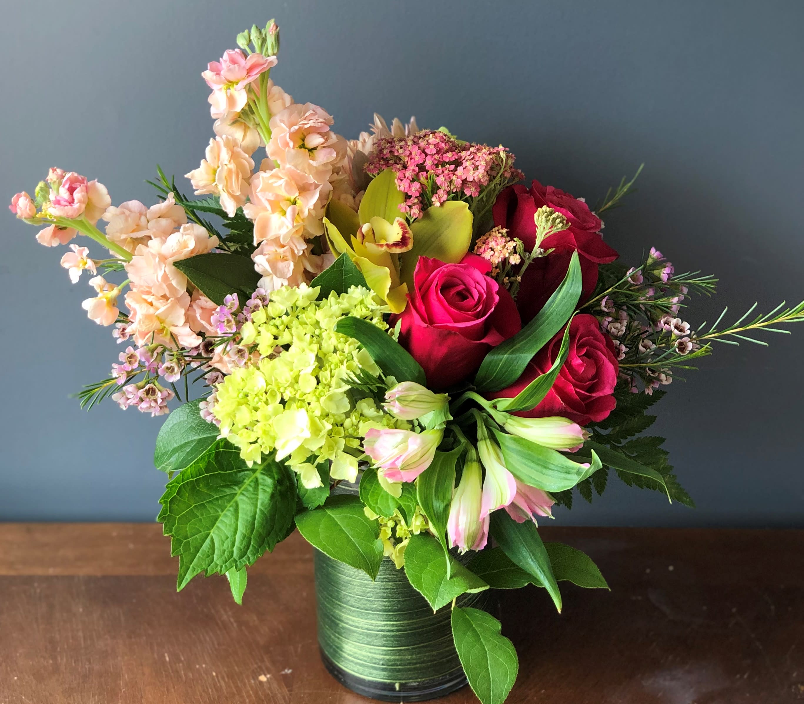 Mixed bouquet of red roses, green hydrangea, orchids, and peach blooms in a green-wrapped glass vase