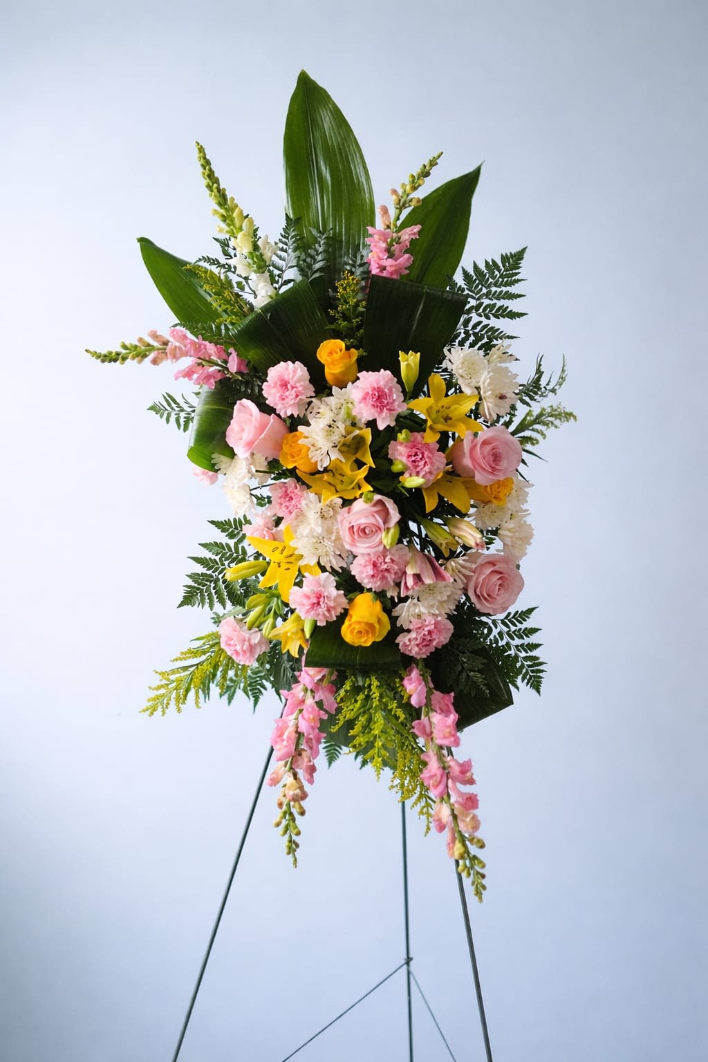 Sunset Serenity Wreath - Soft pink roses and carnations mingle with sunny yellow lilies and roses in a tall standing sympathy arrangement made to comfort and uplift. This funeral spray wreath reads as a gentle, hopeful tribute-the kind of memorial or "I'm sorry" gesture that helps the person you're honoring feel seen. Its pastel and yellow palette brings warmth appropriate for Memorial Day remembrances, celebrations of life, or quiet moments of condolence.  The arrangement combines roses, carnations, lilies, alstroemeria, and vertical snapdragons set within glossy green leaves, airy ferns, and cascading yellow filler for movement and height. Flowers are arranged in an elongated, layered oval that tapers at top and bottom, with long pink snapdragon stems drawing the eye along the full length. Presented on a simple metal easel and slim metal stand, the piece has a polished, formal silhouette ideal for church displays or ceremony spaces.  Choose this wreath when you need a respectful, uplifting tribute for a funeral, memorial service, or to say "I'm sorry" with sincerity; the soft textures and warm colors convey both sympathy and gentle optimism. The standing design arrives ready to display on its metal easel-no assembly needed-and benefits from fresh water in the floral foam until the service; keep it cool and out of direct sun to extend bloom life.