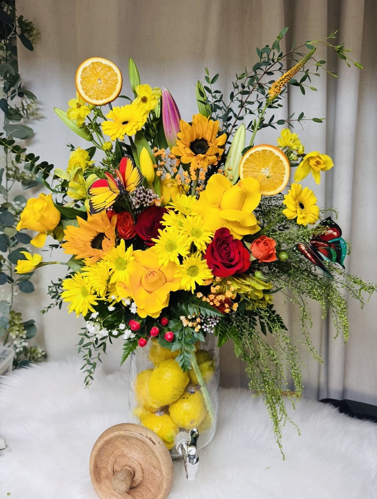 Citrus Sunrise Arrangement - A vibrant and refreshing floral arrangement inspired by the brightness of a summer morning. Featuring cheerful yellow gerberas, vibrant lilies, orange blooms, and red and blush roses, beautifully arranged in a tall clear vase filled with fresh lemons and citrus slices. A stunning choice for birthdays, celebrations, or to add a burst of color to any space approximate height 36”