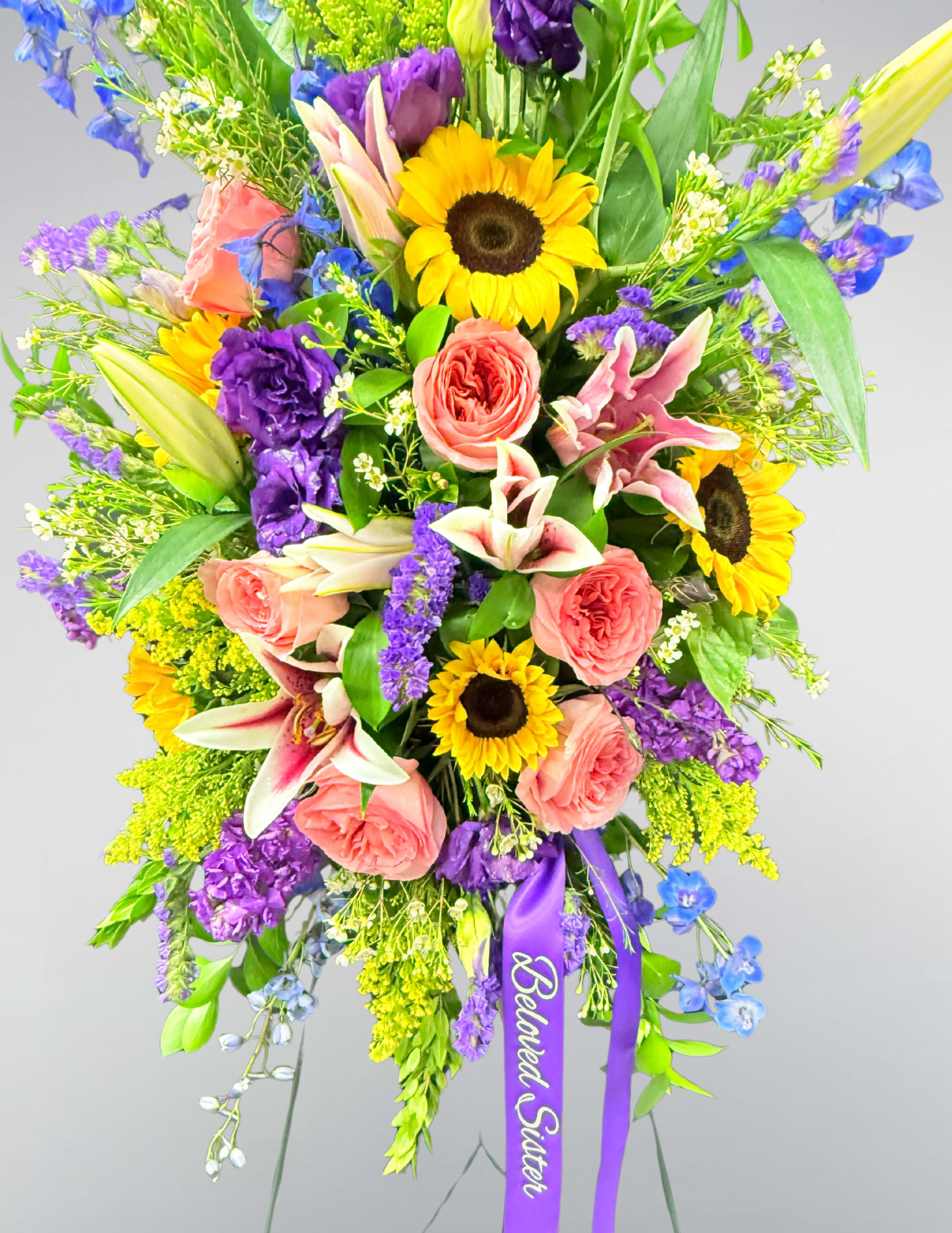 Tall standing floral spray with sunflowers, pink roses, lilies, and purple flowers on an easel
