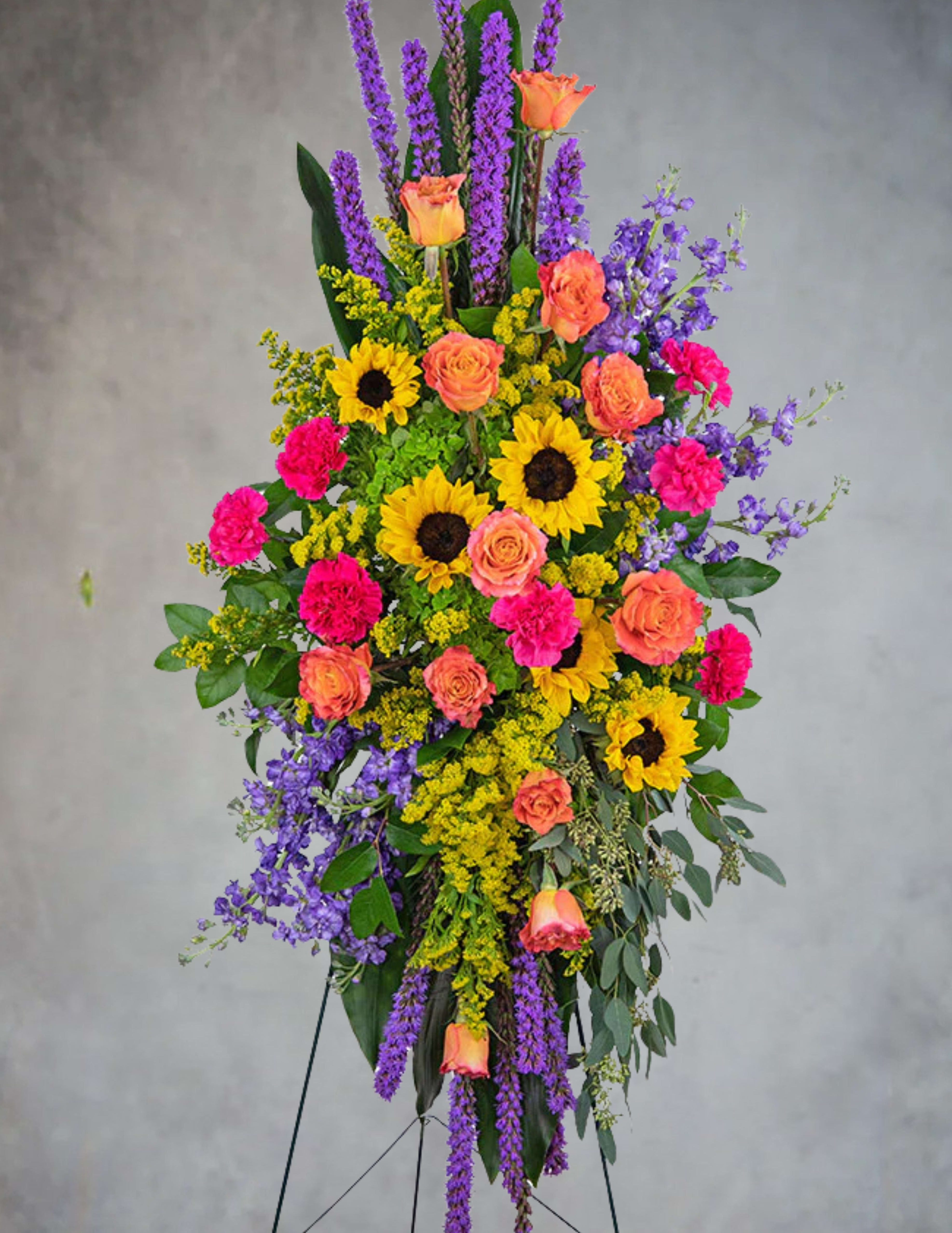 Tall standing spray of sunflowers, orange roses, pink carnations, and purple blooms on an easel.