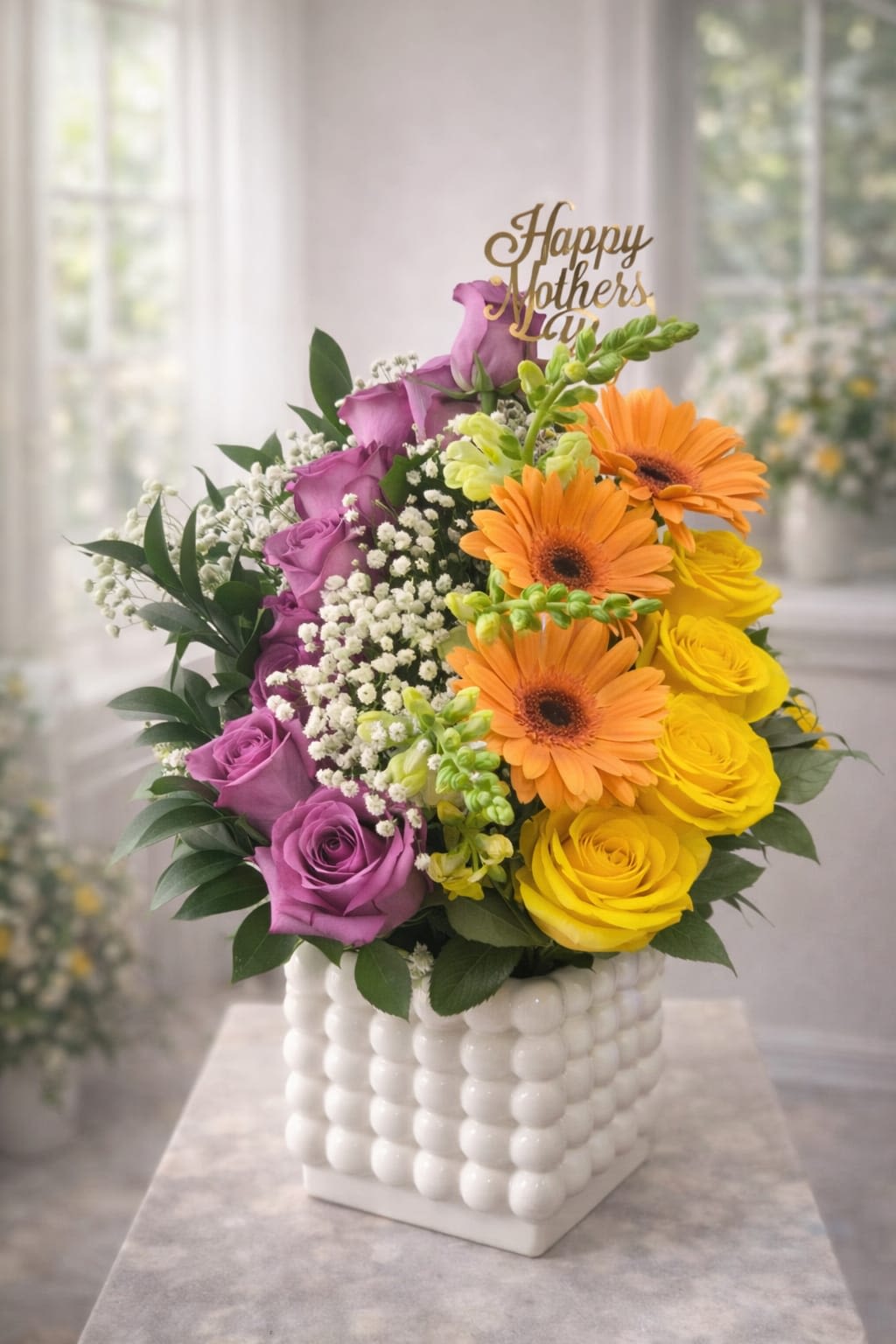 Blooming Love Mother’s Day Arrangement - A vertical sweep of purple roses meets a sunny cluster of yellow roses and bold orange gerbera daisies to create a joyful, modern Mother's Day arrangement that also reads perfectly for birthdays, thank-yous, get-well wishes, or just because. The "Happy Mother's Day" pick gently rises above the bouquet, making the recipient feel celebrated and truly seen while the bright, uplifting palette sends a message of warmth and appreciation.  Soft yellow-green snapdragons and small white filler blooms weave between the larger flowers for airy texture and a light, garden-fresh feel. Arranged in a white textured ceramic cube vase with a raised bubble pattern, the piece has contemporary lines and a tactile finish that suits bedside tables, desks, or a kitchen island. Ideal for the person you want to brighten-mother, mentor, friend, or someone recovering or in need of a cheerful note-this arrangement arrives ready to display and brings an immediate sense of care and delight.