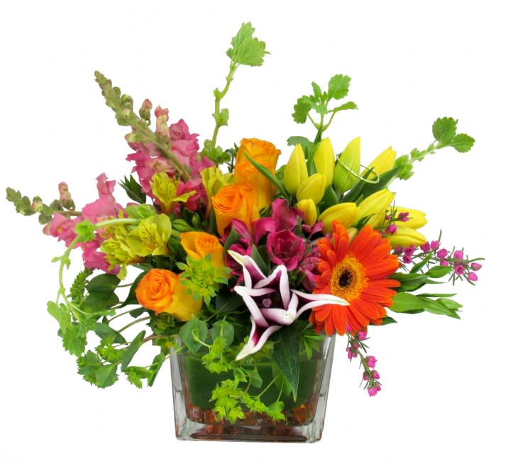 Bright mixed arrangement of orange roses, yellow lilies and assorted blooms in a square glass vase.