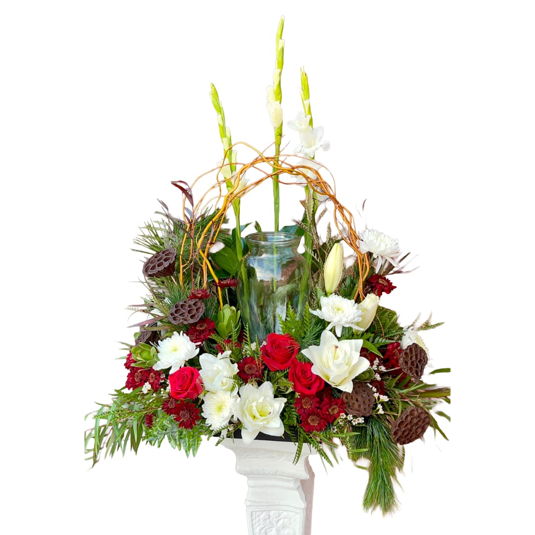 Woodland Rose - A low, circular bed of deep red roses and complementary red mini blooms surrounds a ring of white lilies and chrysanthemums in this sympathy arrangement, creating a heartfelt focal piece for memorial services or a winter holiday gathering. Evergreen textures and looping curly branches give the design a lush, layered feel while tall white flower spikes lift the composition-making the person you're honoring feel seen and remembered.  Dried lotus pods punctuate the palette with rustic, sculptural brown accents, and a central clear glass cylinder sits ready to hold a candle or a small urn, framed by the flowers and vertical movement. The arrangement is presented on a white pedestal stand for formal display; dimensions are low and circular with taller central elements, ideal when you need a respectful centerpiece that reads well from all sides.  Designed for sympathy tributes, memorial services, or as a solemn holiday display, this piece communicates warmth, reverence, and quiet beauty to the recipient's family and friends. Minimal care is needed for delivery-keep the glass cylinder clean if used for a candle, and place away from drafts to preserve the lilies and chrysanthemums during events.
