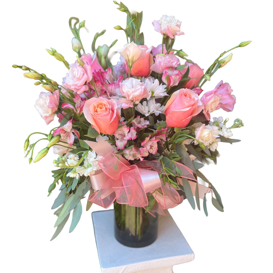 Pretty in Peach - A soft, pastel bouquet that reads like a garden in miniature-pink and peach roses mingle with blush lisianthus, pale stock and sprightly alstroemeria to create a rounded birthday bouquet or anniversary arrangement that feels intimate and thoughtfully made. The first impression is romantic and comforting: the person you're thinking of will feel gently celebrated the moment they see the delicate color story and the large pink organza bow that crowns the design.  Flowers are densely clustered at the center and taper outward for a full, textured silhouette, with white and pink accent blooms adding light and movement. Stems are arranged in a clear glass cylinder vase so the clean lines of the glass and the visible stems balance the soft, garden-like top. Finished on a white display pedestal, this arrangement is an elegant choice for Mother's Day, a get-well wish, or simply to say "I'm thinking of you" with warmth and grace.