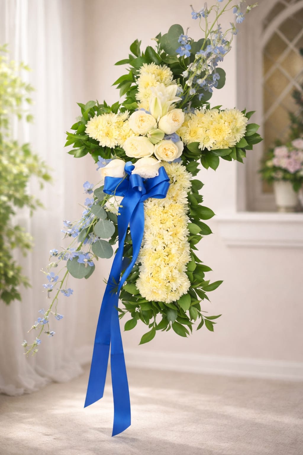 Heavenly Peace Cross - A gently reverent cross of pale yellow chrysanthemums creates the first, calming impression-perfect for funeral services, Memorial Day remembrances, or an Easter tribute. This sympathy arrangement in the shape of a cross conveys quiet comfort and faith, the kind of floral expression that lets the person you're honoring feel remembered and tenderly held.  Creamy white roses and a single white lily cluster near the center provide soft focal points, their rounded and trumpet blooms contrasting the chrysanthemum texture. Light blue delphinium and small blue accents trail along one side, picked up by a vivid royal blue satin ribbon with long tails at the center; a collar of deep green foliage frames the silhouette for a clean, dignified outline. Sized to serve as a focal spray for services or a prominent memorial display, it reads as both formal and deeply personal.  Send this arrangement when words fall short-whether you're saying I'm sorry, offering thanks, or simply thinking of someone during a difficult time. For display guidance and delivery timing for services or graveside placement, select the appropriate delivery date at checkout so the cross arrives fresh and ready for viewing.