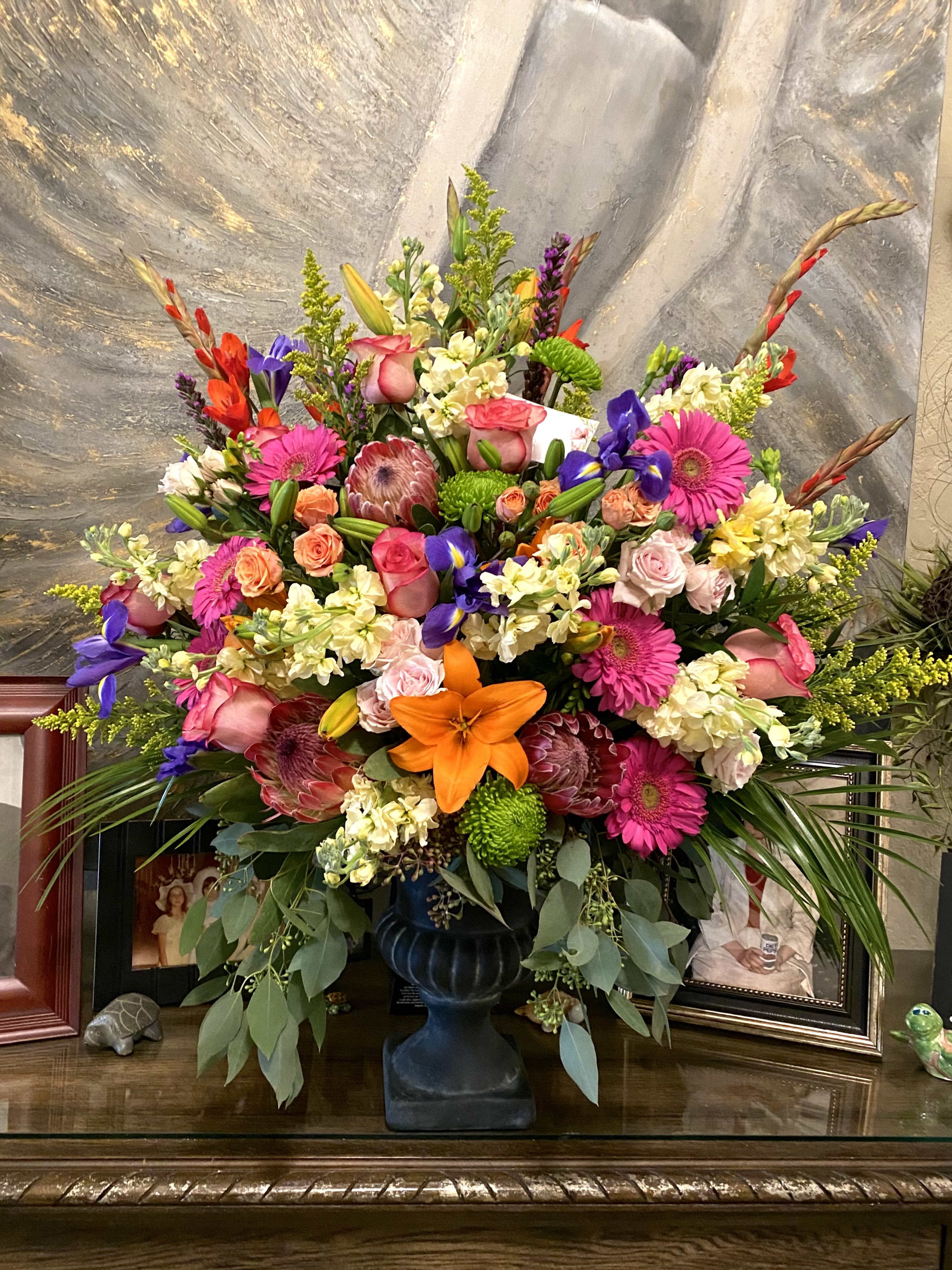 Majestic Garden - A towering, fan-shaped display that reads like a walk through a vibrant garden-this Majestic Garden flower arrangement makes an immediate statement for a birthday bouquet, anniversary roses alternative, or an elegant sympathy arrangement. The mix of pink and peach roses with bright pink gerbera daisies and vivid orange lilies creates a joyful yet refined center, while the purple iris and red-orange snapdragon spikes add vertical drama that draws the eye upward.  Rich textures and unexpected accents give the arrangement depth: protea and green button-style chrysanthemums nestle among creamy clusters of smaller blooms, and the overall palette of pink, orange, purple, and white keeps the design lively and celebratory. The flowers are arranged in a dark pedestal urn vase for a formal silhouette, with optional keepsakes-picture frames and a small turtle figurine-available to personalize the display and make it feel truly thoughtful.  Perfect for honoring milestones or offering comfort, Majestic Garden is sized to serve as a focal point in a living room, entryway, or memorial table; its height and fullness ensure it reads beautifully from across a room. Standard care-fresh water, a cool spot out of direct sun, and trimming stems if rearranged-will help the blooms last and keep the presentation as striking as the moment you give it.