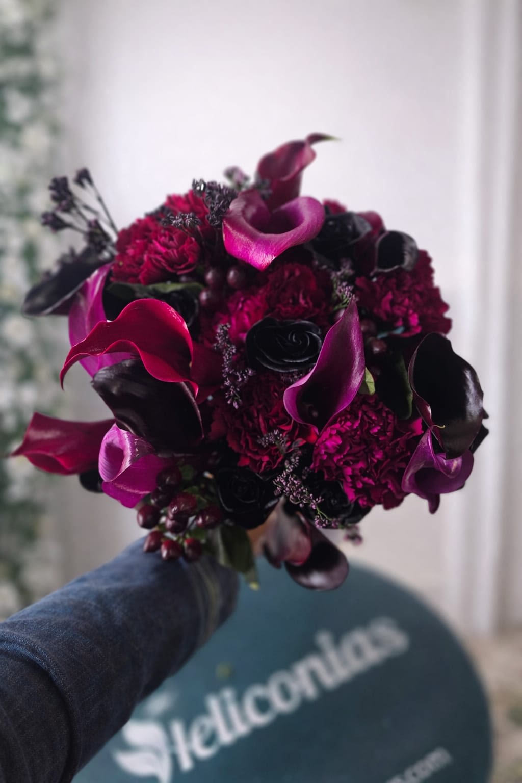 Midnight Romance Bridal Bouquet - A dense cluster of deep berry and crimson blooms sets a moody, romantic tone-perfect as a wedding bouquet for an evening ceremony or a bride who wants bold, dramatic florals. This bridal bouquet for weddings combines sculptural magenta mini calla lilies and ruffled red carnations with very dark roses and inky berries, creating striking contrast and a sophisticated, romantic silhouette that will make the person you're celebrating feel utterly seen.  Hand-tied for a natural, easy-to-carry look, the arrangement pairs glossy calla curves with textured carnation heads and near-black roses that act as dramatic highlights. The palette of pink, red, purple and black reads rich and velvety in photos and candlelight; its compact, dense form makes it ideal for close-up portraits and sweeping evening vows.