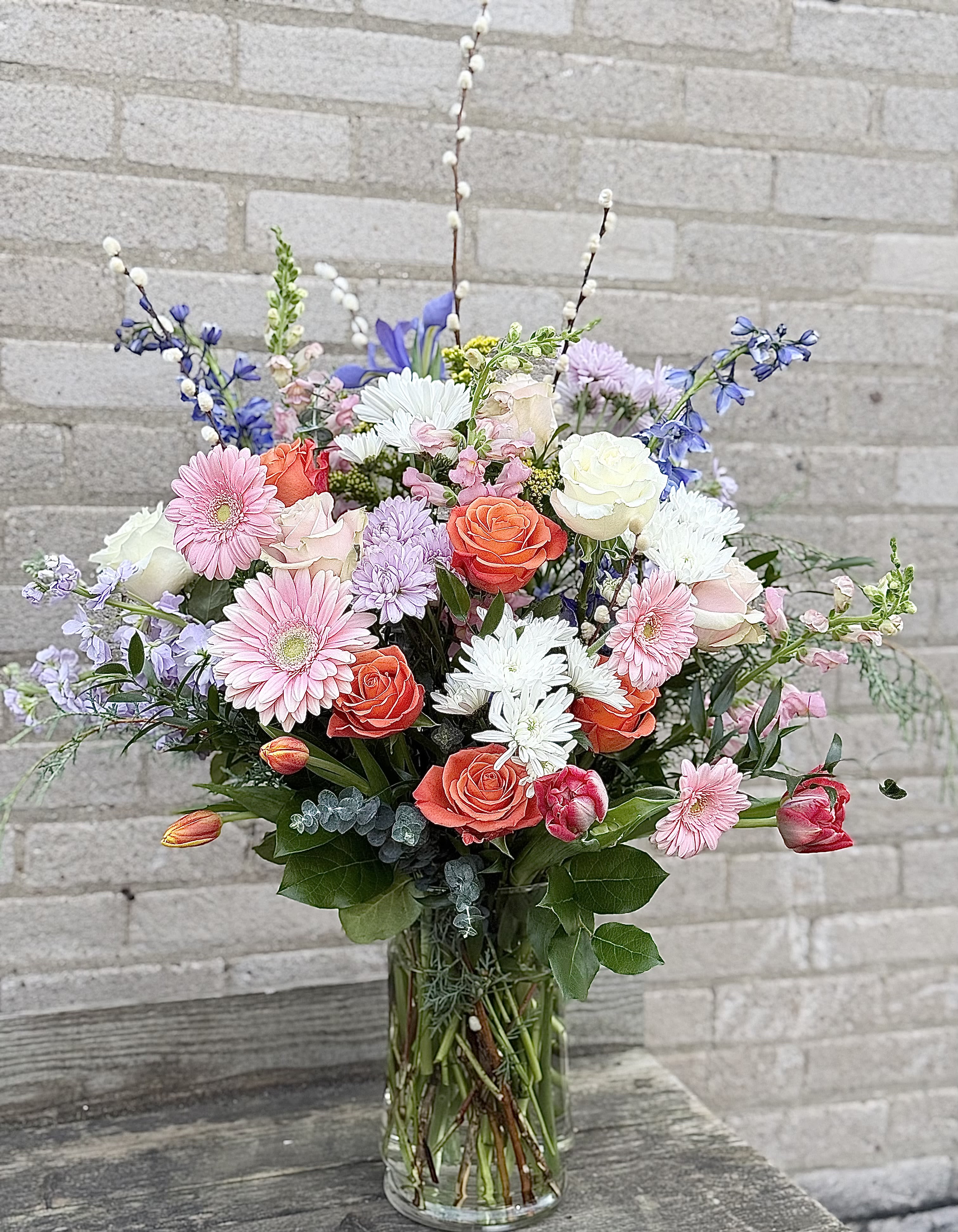 Grand Color Expression - A tall, airy bouquet that greets the eye with pink gerbera daisies and bright orange roses-perfect as a birthday bouquet, Mother's Day arrangement, or an anniversary surprise. The Grand Color Expression (arrangement) creates a garden-like silhouette that makes the person you're thinking of feel celebrated: cheerful, elegant, and uplifted without feeling formal.  This flower arrangement pairs white and lavender chrysanthemums and soft pastel filler blooms with sky-blue delphinium spikes and upright snapdragon stems for vertical interest. Tulips and mixed greenery soften the composition while a clear glass cylinder vase keeps the look fresh and modern; overall the design reads light and spring-ready but works year-round. Ideal for sending get-well wishes, congratulations, or a warm thinking-of-you message-sizes fit standard gift needs and arrive arranged and ready to display with simple care: fresh water, a cool spot away from direct sun, and a daily water refresh to prolong bloom life.