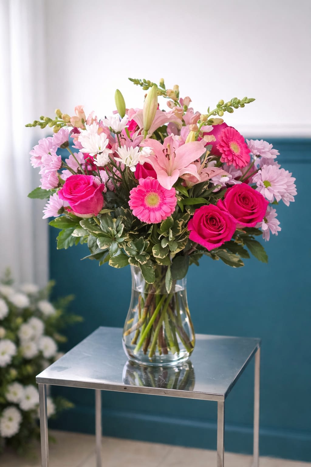 Blushing Fuchsia Elegance Vase - Vivid pink roses and gerbera daisies meet pale pink lilies and soft lavender chrysanthemums in a rounded, full bouquet that reads like a joyful embrace - ideal as a birthday bouquet, Mother's Day arrangement, anniversary flowers, or a bright get-well gift. The mix of hot pinks with gentle pastels gives the recipient an immediate lift; taller lily buds and snapdragon stems add graceful height so the bouquet reads as both abundant and elegant.  Arranged in a clear glass vase, the blooms form a compact, sculpted shape with textural contrast between the velvety roses, the bold daisy faces, and the feathery chrysanthemum clusters. The palette of pink, white, lavender, and mixed tones looks fresh against contemporary settings - pictured here on a sleek metal side table - making it a fitting centerpiece for a dining table, bedside, or office desk.  Choose this arrangement to make someone feel celebrated and seen: it sends a warm, affectionate message without being overdone. The vase is included for easy display; lilies may open over a few days for added drama - trim stems and refresh water regularly to prolong bloom life.