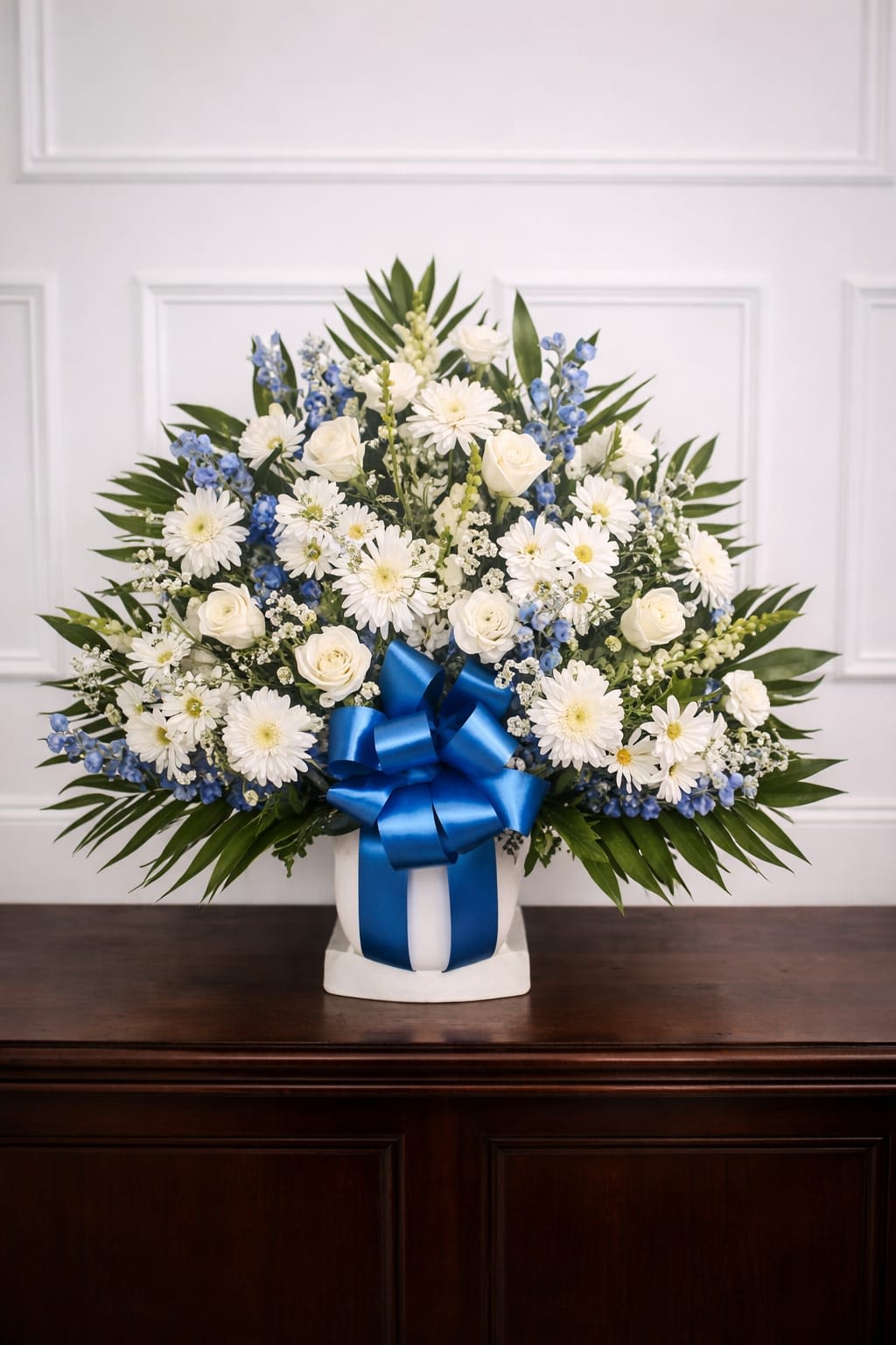 Peaceful Tribute Altar Arrangement - A fan-shaped display of white and blue flowers creates a calm, dignified presence ideal for sympathy and funeral settings or for saying "I'm sorry" and "thinking of you." This altar arrangement is built in a white urn on a matching pedestal base so it reads formal and respectful; the large blue satin bow at the front gives a gentle, consoling accent that feels appropriate for Memorial Day remembrances and year-round memorial moments.  Clusters of white roses and daisy-style chrysanthemums are punctuated by vertical white snapdragons and tall blue delphinium, with elongated green foliage framing the outer edges to form a broad, structured silhouette. The combination of textures-soft rose petals, feathery chrysanthemum centers, and the upright strength of delphinium-creates visual depth while keeping the palette cool and serene.  Designed to honor and comfort the person you're remembering, this arrangement suits memorial services, hospital rooms for get-well wishes, or a quiet expression of gratitude and condolence. The arrangement arrives ready in its white urn and pedestal; keep blooms cool and away from direct heat to extend their presence during services and viewings.