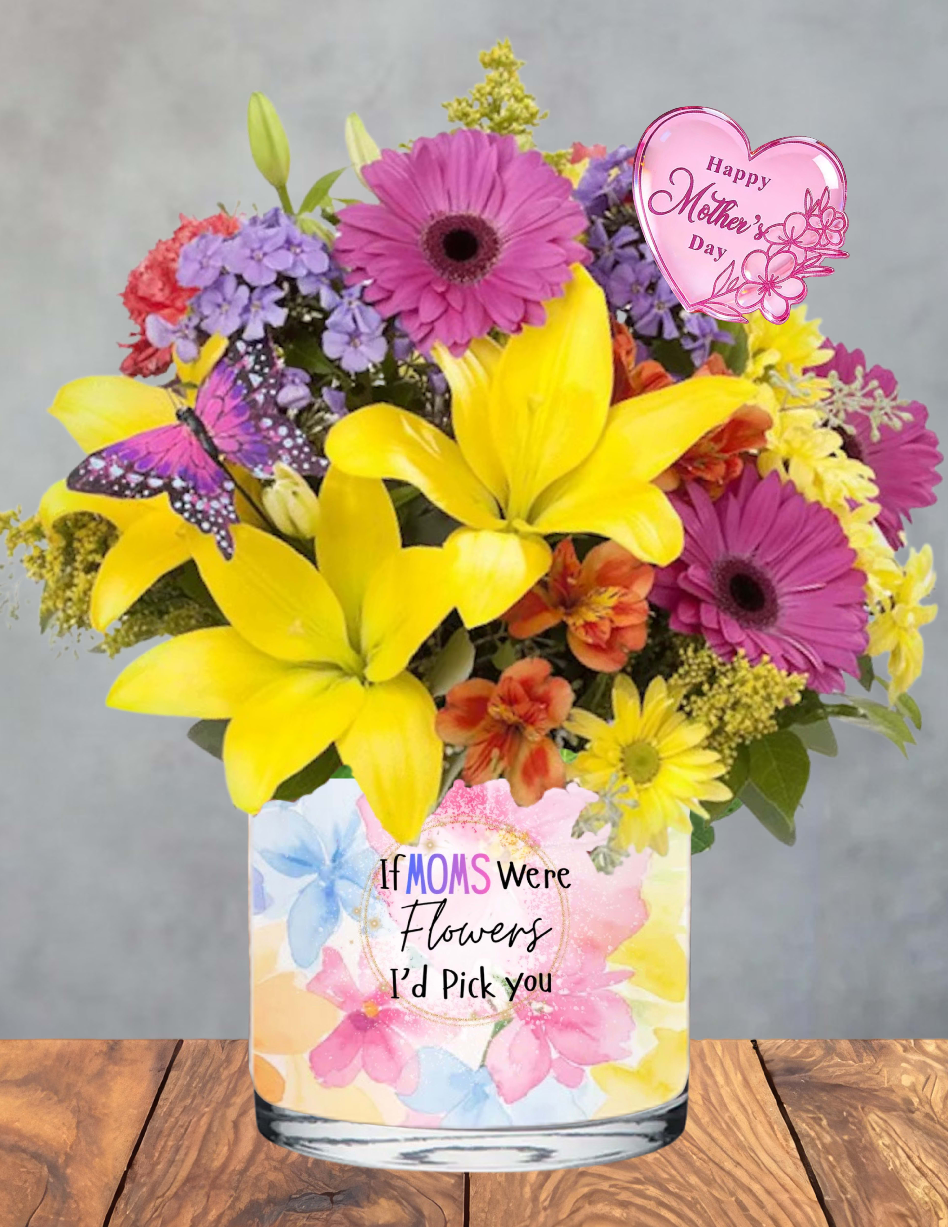 Mother’s Day Radiance - Mother’s Day Radiance A bright, heart‑lifting celebration of everything Mom brings into the world.  Mother’s Day Radiance glows with sun‑kissed lilies, vibrant gerbera daisies, rich red and orange blooms, and soft touches of purple and pink, all gathered in a lush, cheerful embrace. A delicate butterfly rests among the petals, adding a whisper of whimsy and grace.  Arranged in a charming floral vase adorned with the sentiment “If Moms Were Flowers, I’d Pick You,” this bouquet shines with gratitude, warmth, and love. It’s the perfect way to honor her light, her strength, and the beauty she brings to every day.