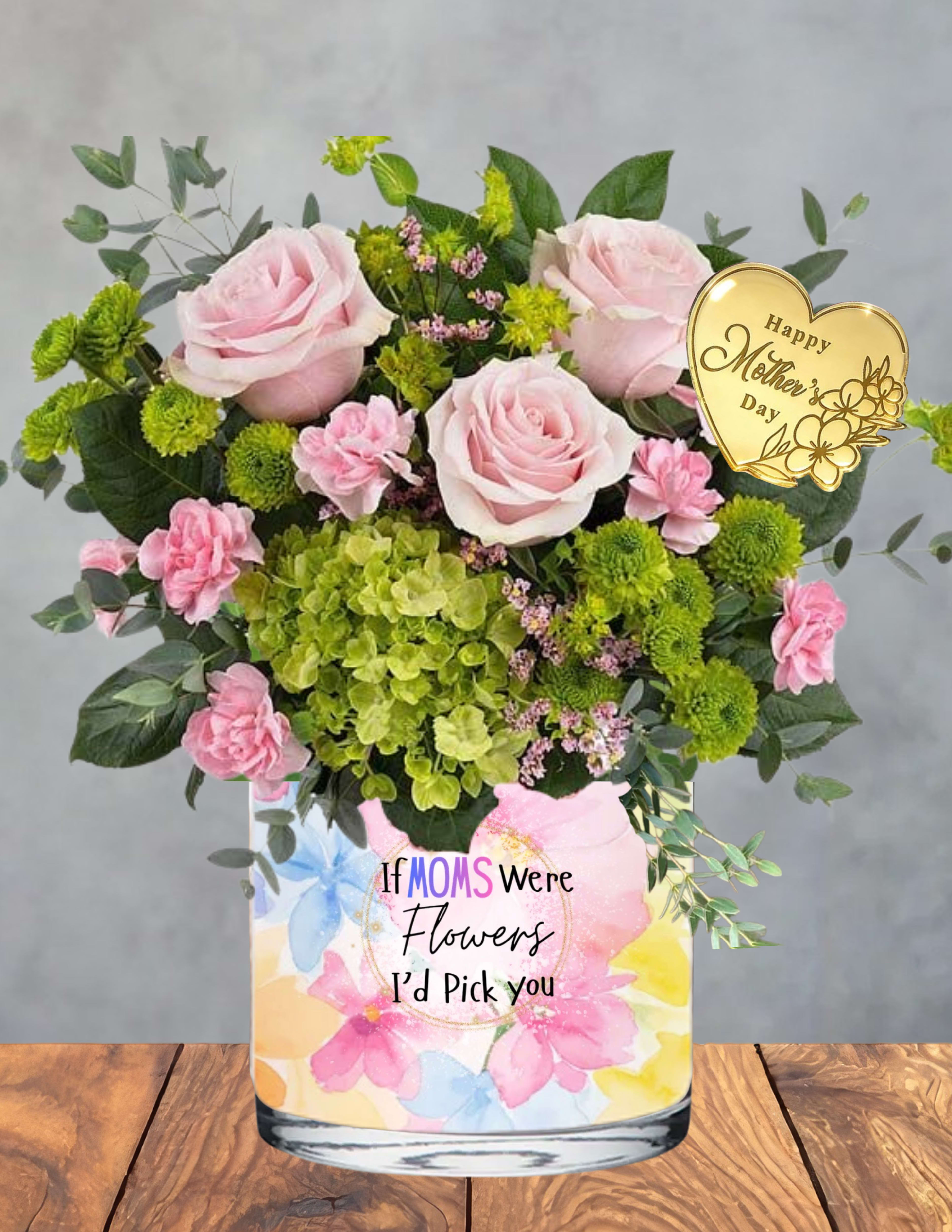 Moms Serene Meadow - A soft, peaceful garden moment crafted just for her.  Mom’s Serene Meadow brings together blush‑pink roses, petite carnations, and fresh green hydrangeas, creating a soothing blend of color and texture that feels like a quiet breath of spring. Green button mums and lush foliage add natural depth, giving the arrangement its meadow‑like calm and effortless beauty.  Tucked among the blooms is a gold heart keepsake engraved with a loving Mother’s Day message — a warm, elegant touch she can treasure long after the flowers have faded. Presented in a pastel floral vase with the sentiment “If Moms Were Flowers, I’d Pick You,” this design radiates gratitude, tenderness, and the gentle joy of being loved.  A perfect choice for the mom who brings peace, grace, and steady warmth to every day.