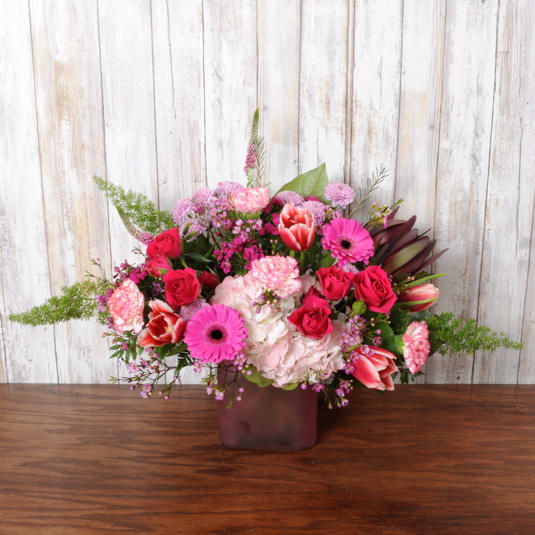 Bright pink and red mixed flower arrangement in a frosted cube vase on a wooden table.