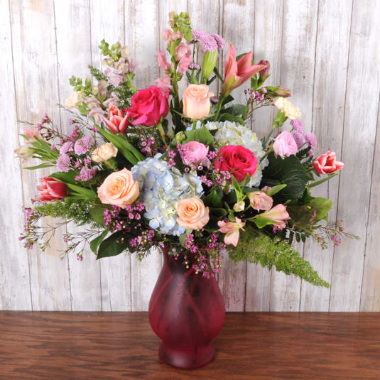 Tall mixed arrangement of pink roses, lilies, tulips, and blue hydrangeas in a red glass vase