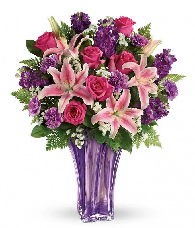 Pink lilies, roses, and purple flowers arranged in a tall purple glass vase
