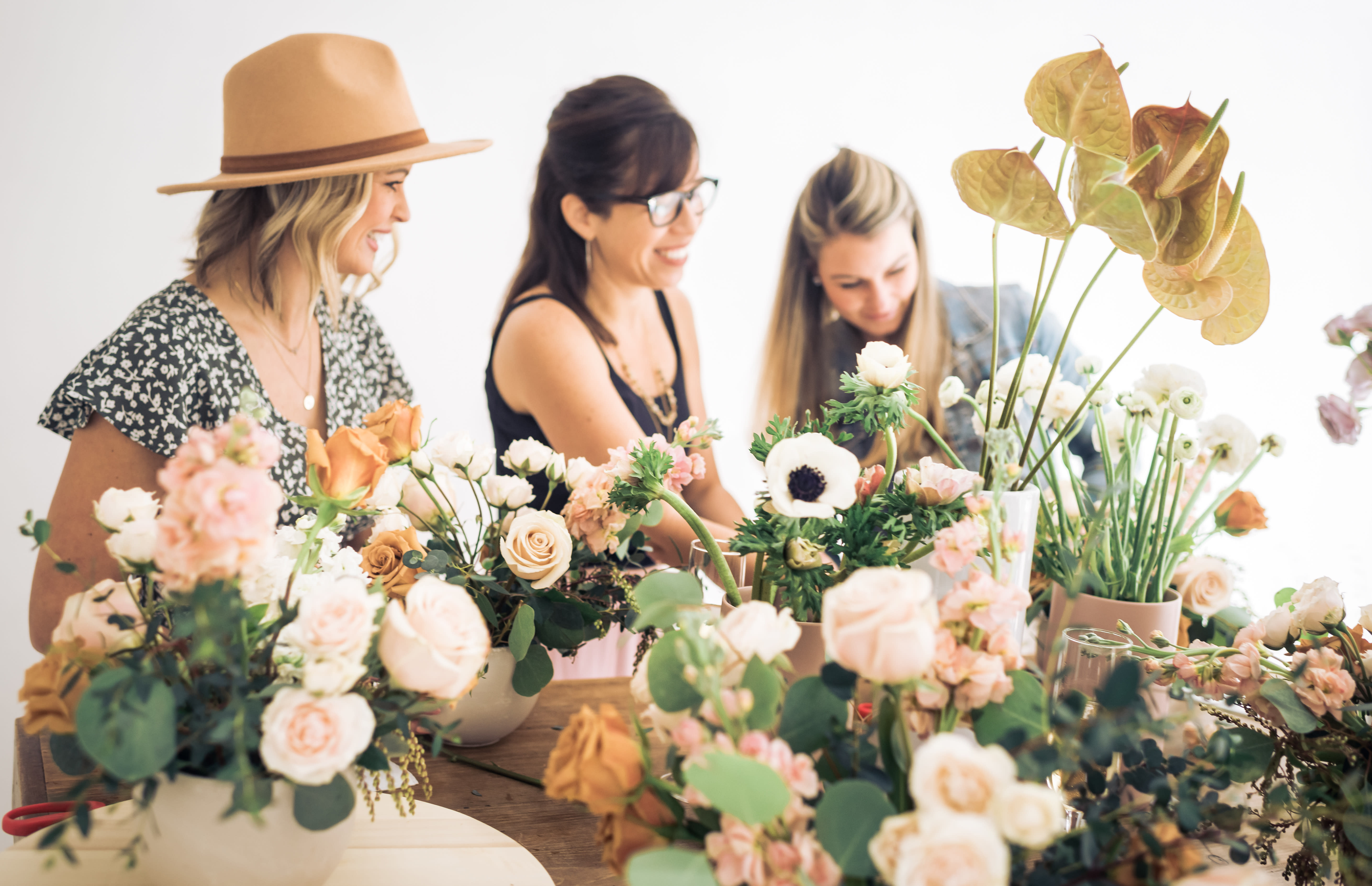 Three women arranging blush and white flower centerpieces on a wooden table