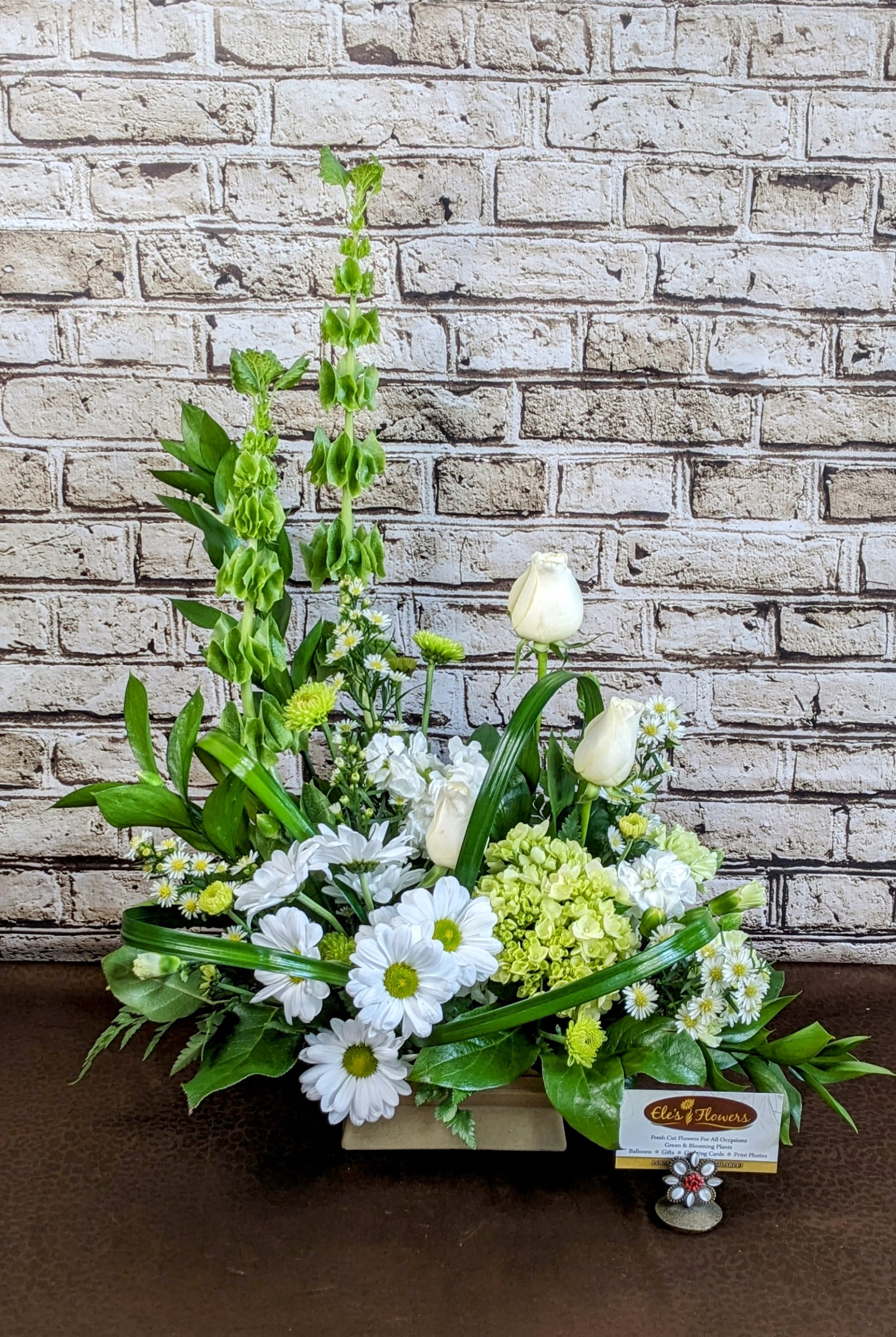 Rectangular arrangement of white daisies, white roses, green hydrangea, and tall green Bells of Ireland.