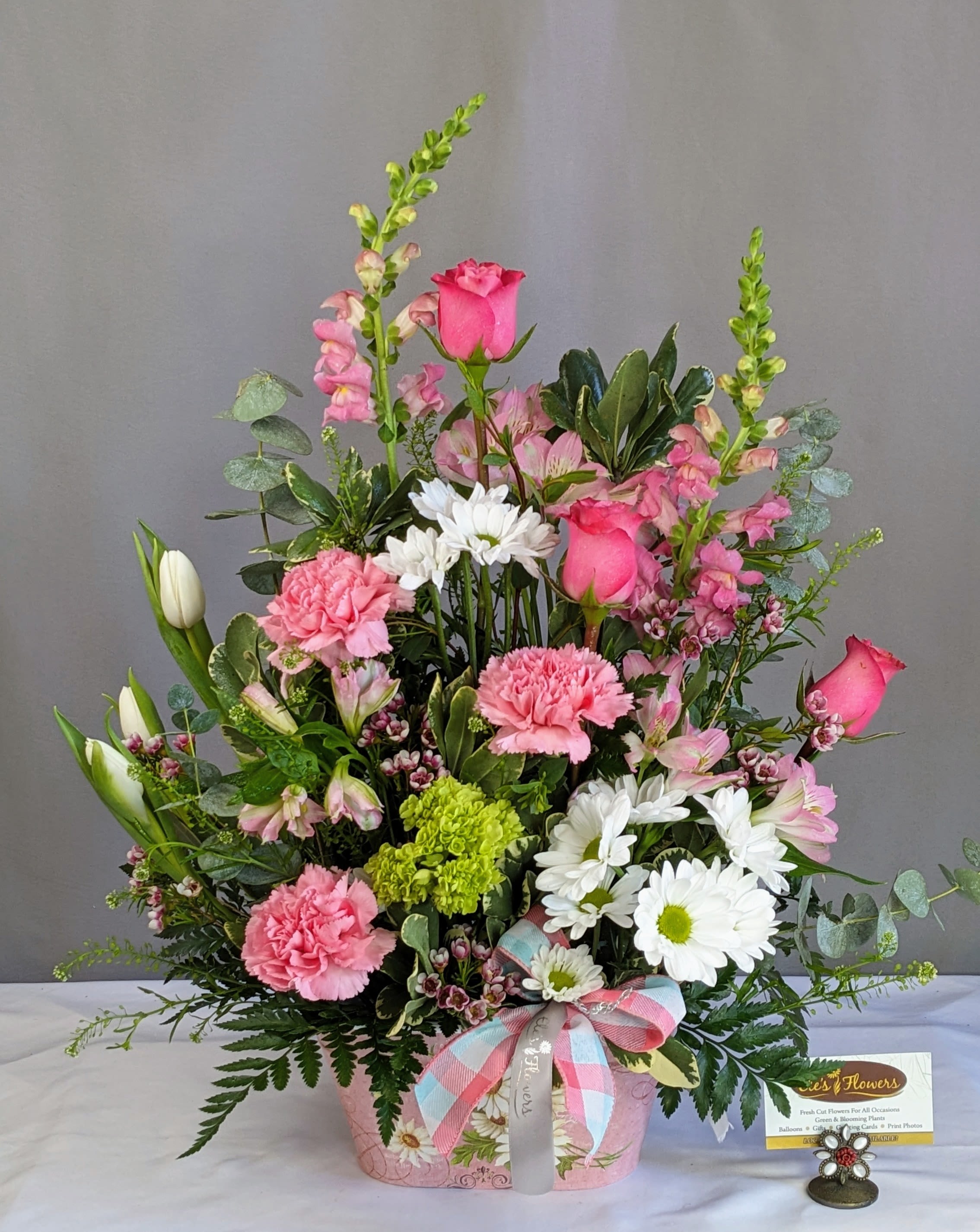 Gentle Grace Tribute  - This heartfelt sympathy arrangement offers a gentle expression of comfort and remembrance, designed in a soft pastel palette that brings a sense of peace and warmth. A harmonious blend of delicate pinks, crisp whites, and fresh greens creates a soothing, uplifting presence—perfect for honoring a loved one’s memory.  Arranged in a charming keepsake container adorned with a soft, coordinating ribbon, this design features a lush mix of textures and heights, giving it a natural, garden-inspired feel. Graceful accents rise above the arrangement, while full, rounded blooms and airy greenery fill in below, creating a balanced and comforting display.  Thoughtfully designed to convey love, support, and quiet strength, this arrangement is suitable for funeral services, visitations, or as a meaningful gesture sent to the home.