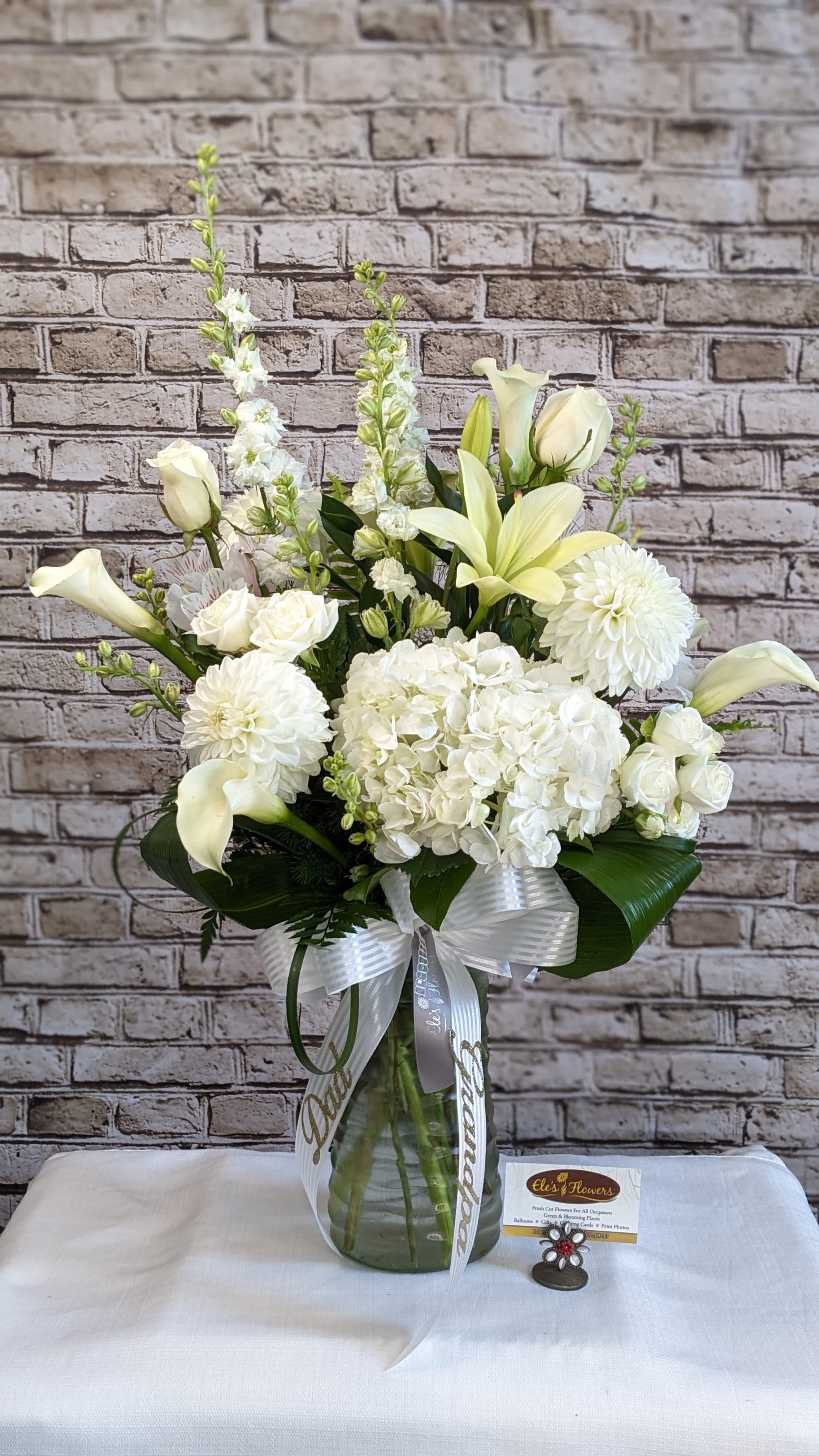 Tall white floral arrangement with lilies, roses, hydrangeas and calla lilies in a glass vase with white ribbon