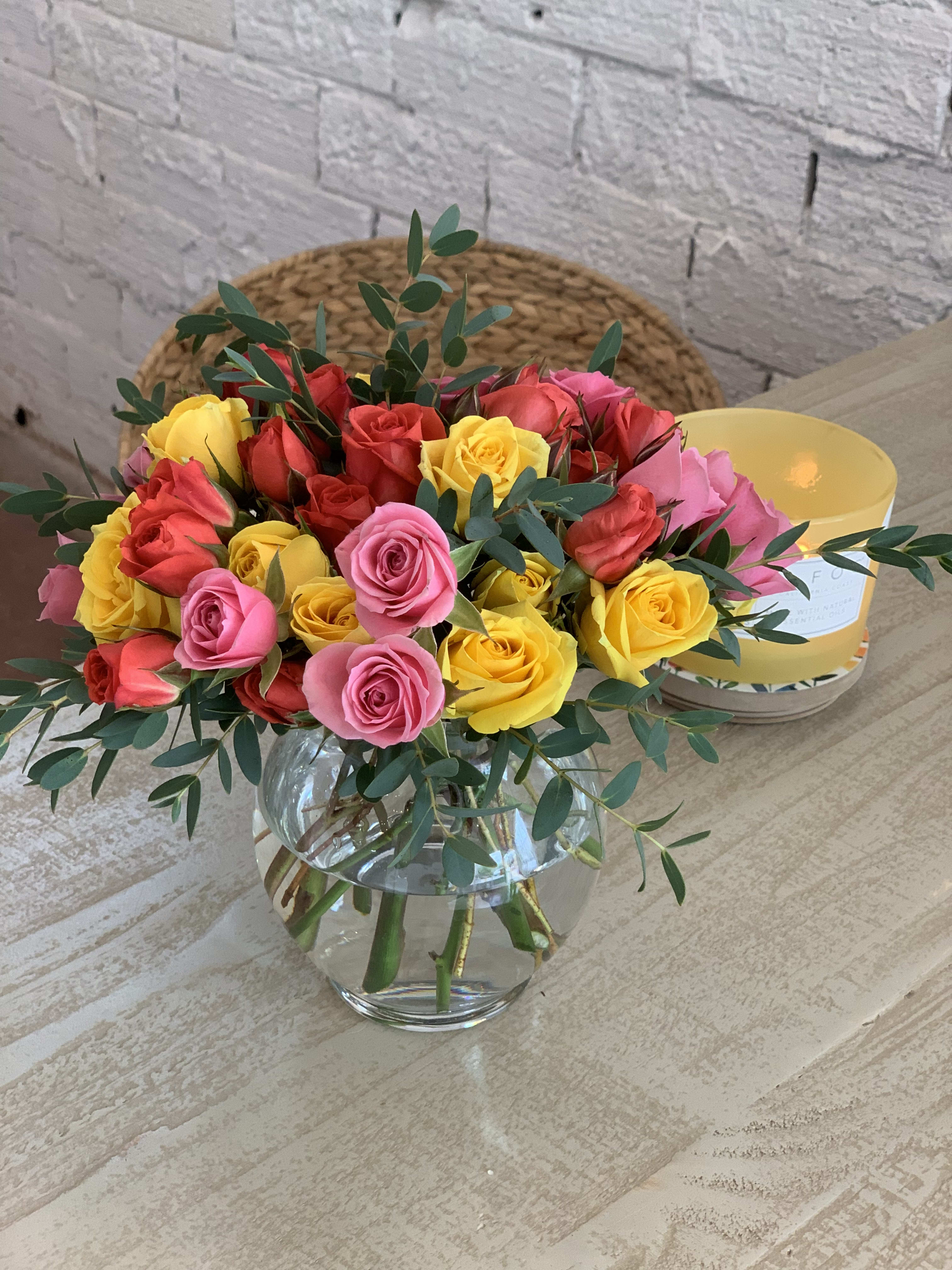 Round glass vase of red, pink, and yellow roses accented with eucalyptus on a light wood table.